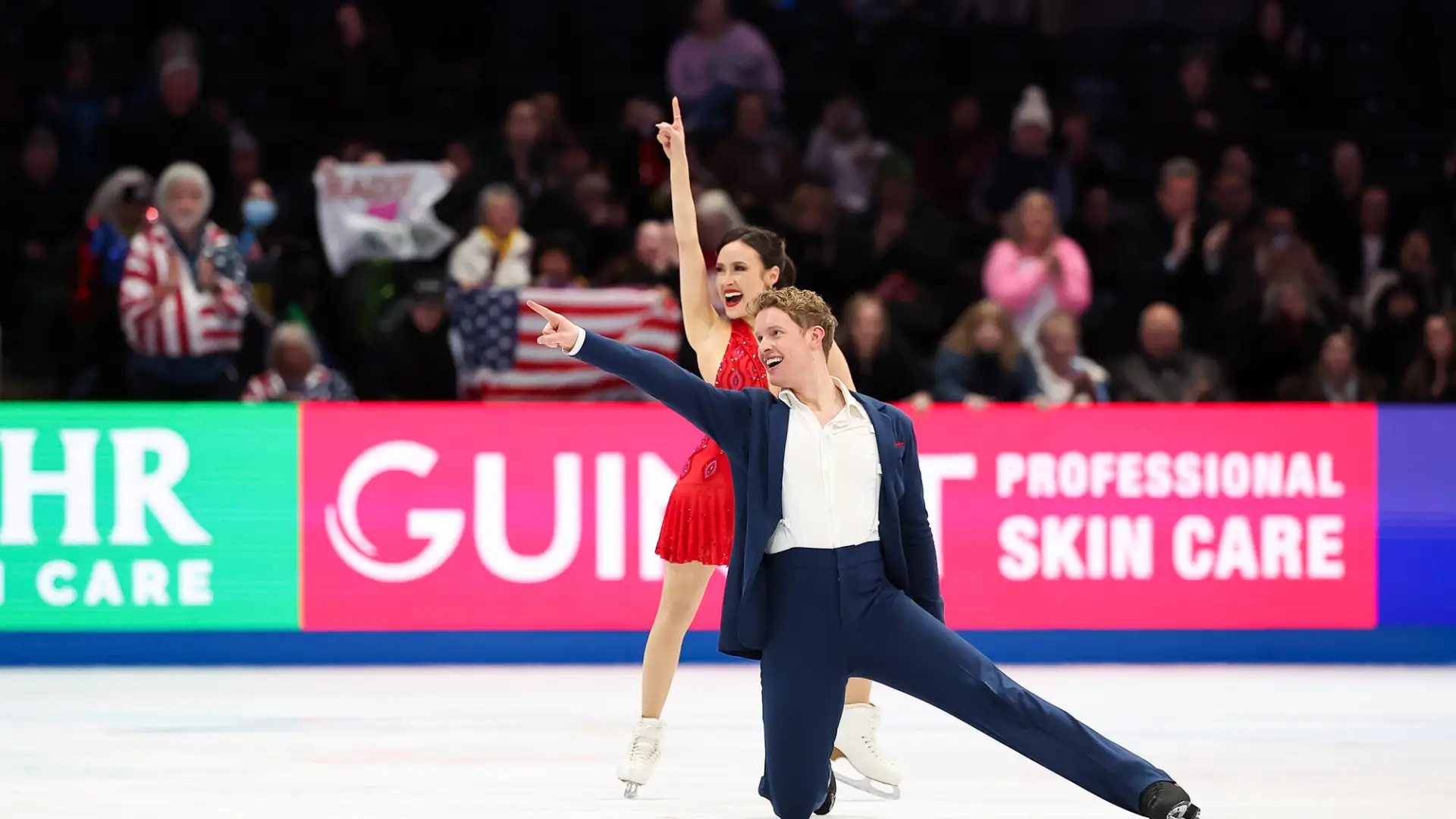 Male and female figure skating pair perform in blue suit and red dress respectively
