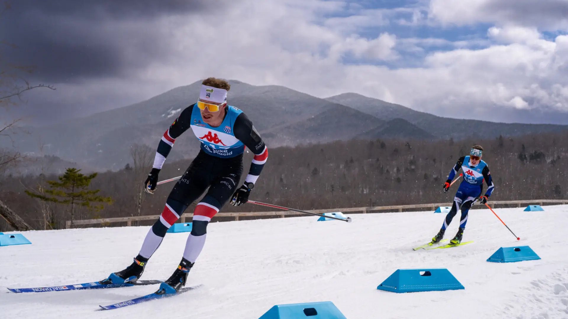 The image shows two cross-country skiers racing on a snowy course. 