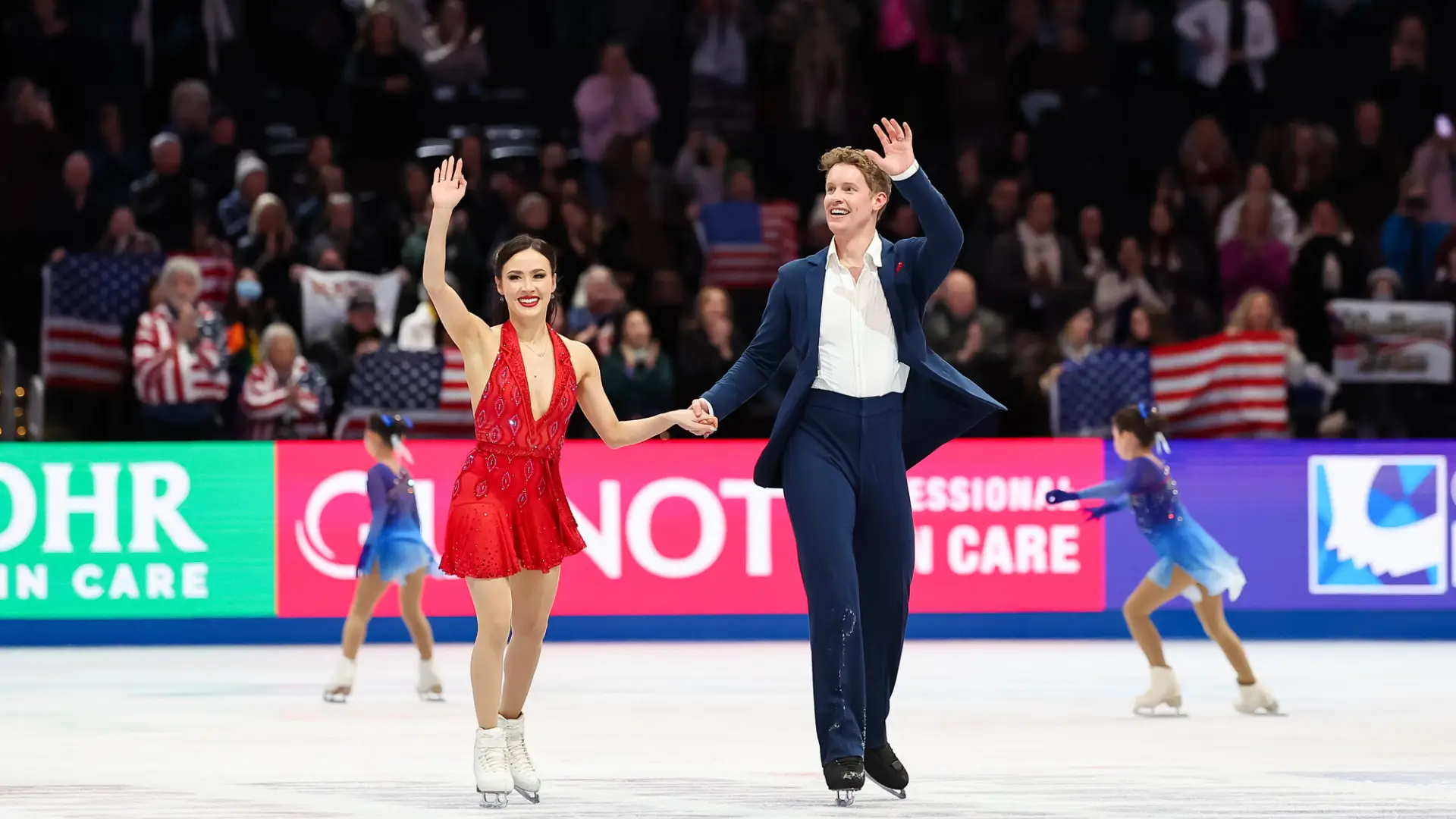 Pair of figure skaters, a woman in a red dress and a man in a blue suit, wave to crowds from on the ice while young girls in blue dresses skate behind