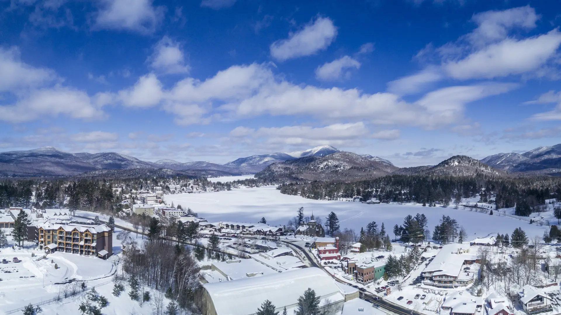 Aerial photo of the Village of Lake Placid in winter. Mirror Lake is covered in snow, as are all the buildings of downtown and the mountains in the distant background. 