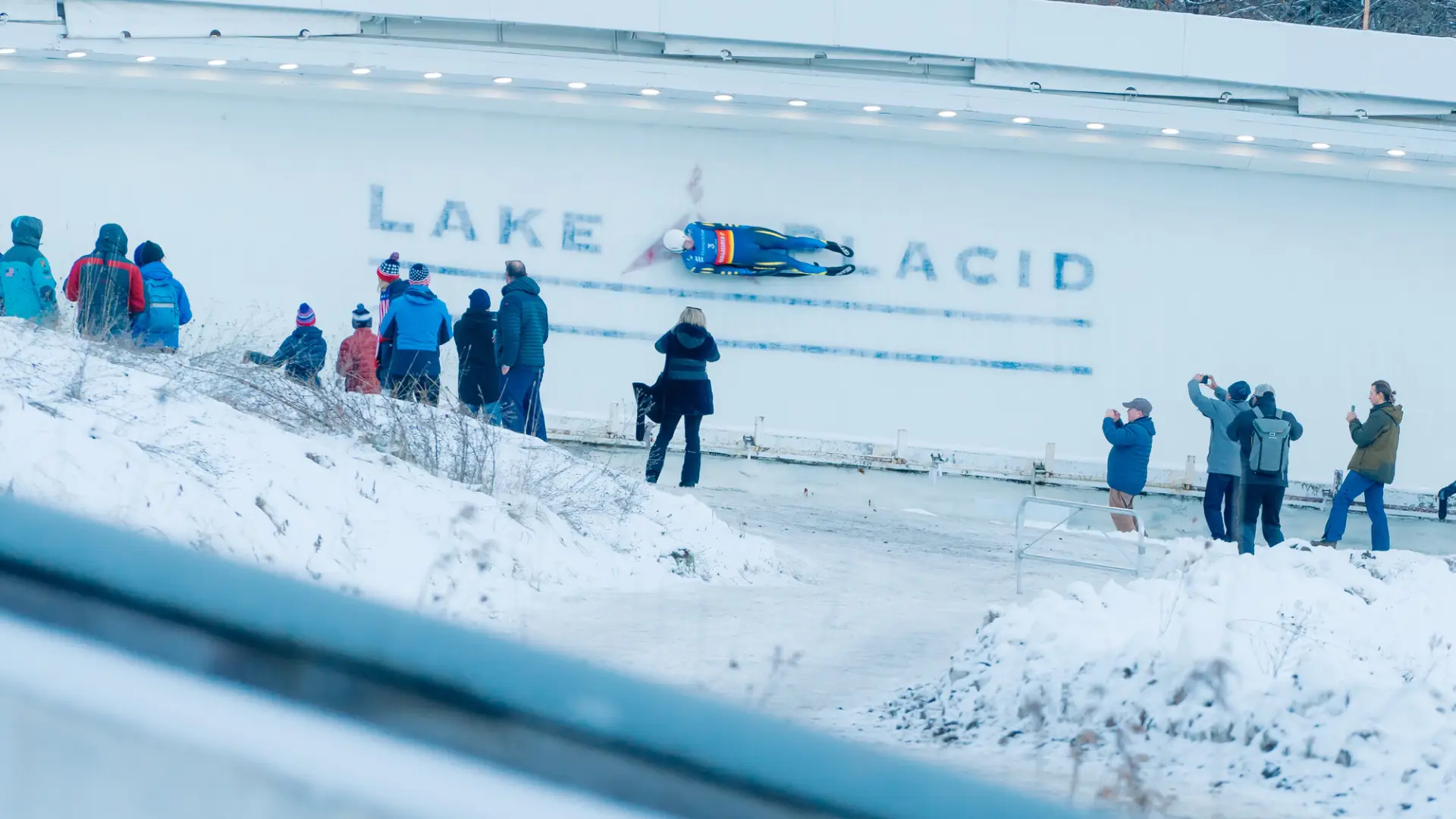 Luge athlete rides down track over where Lake Placid is printed into the ice while several people look on, some taking photos