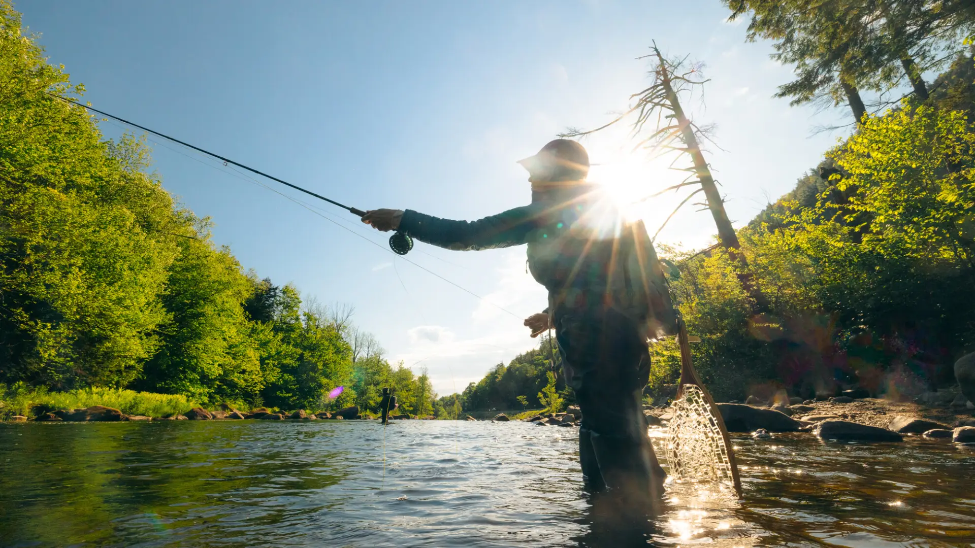 A fly fisherman casting a line in a river on a sunny day.