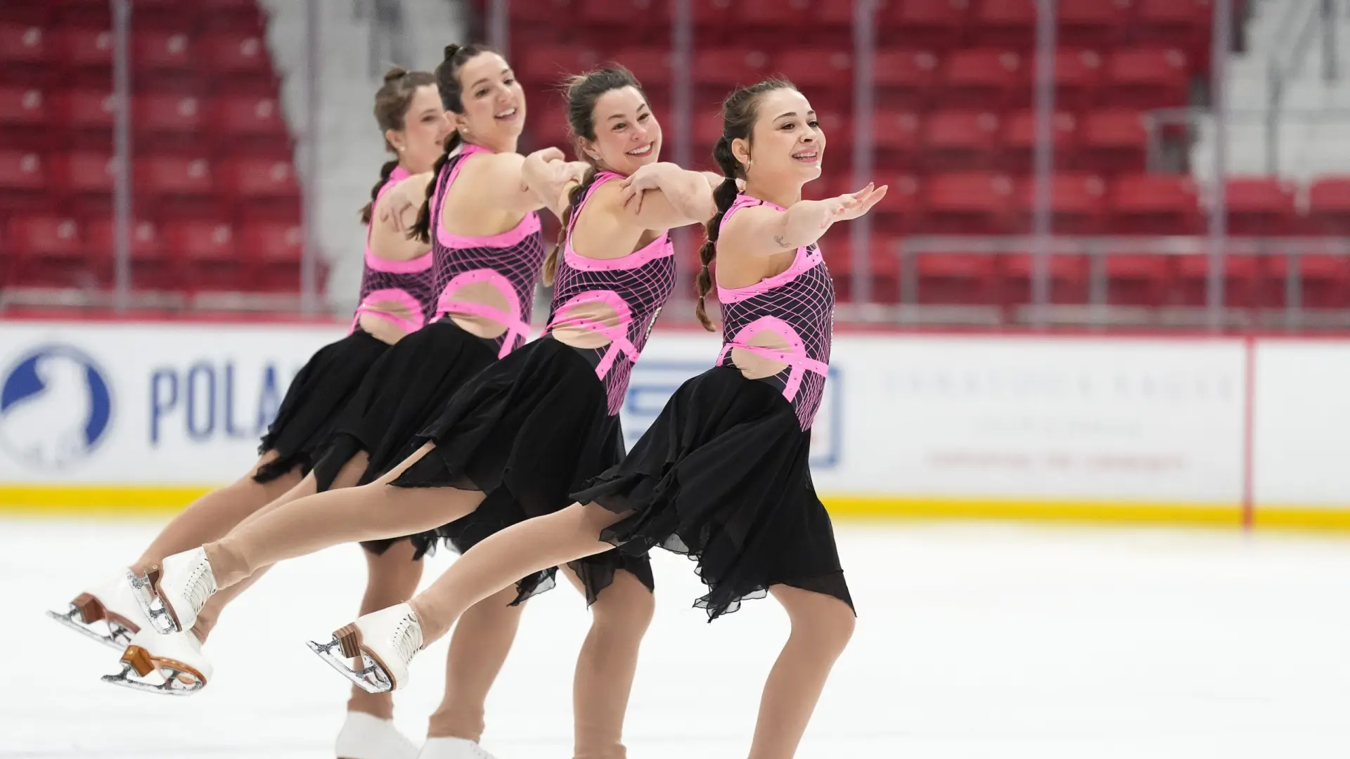 A synchronized ice skating team performs a routine in matching pink and black outfits, showcasing graceful movements on the rink.