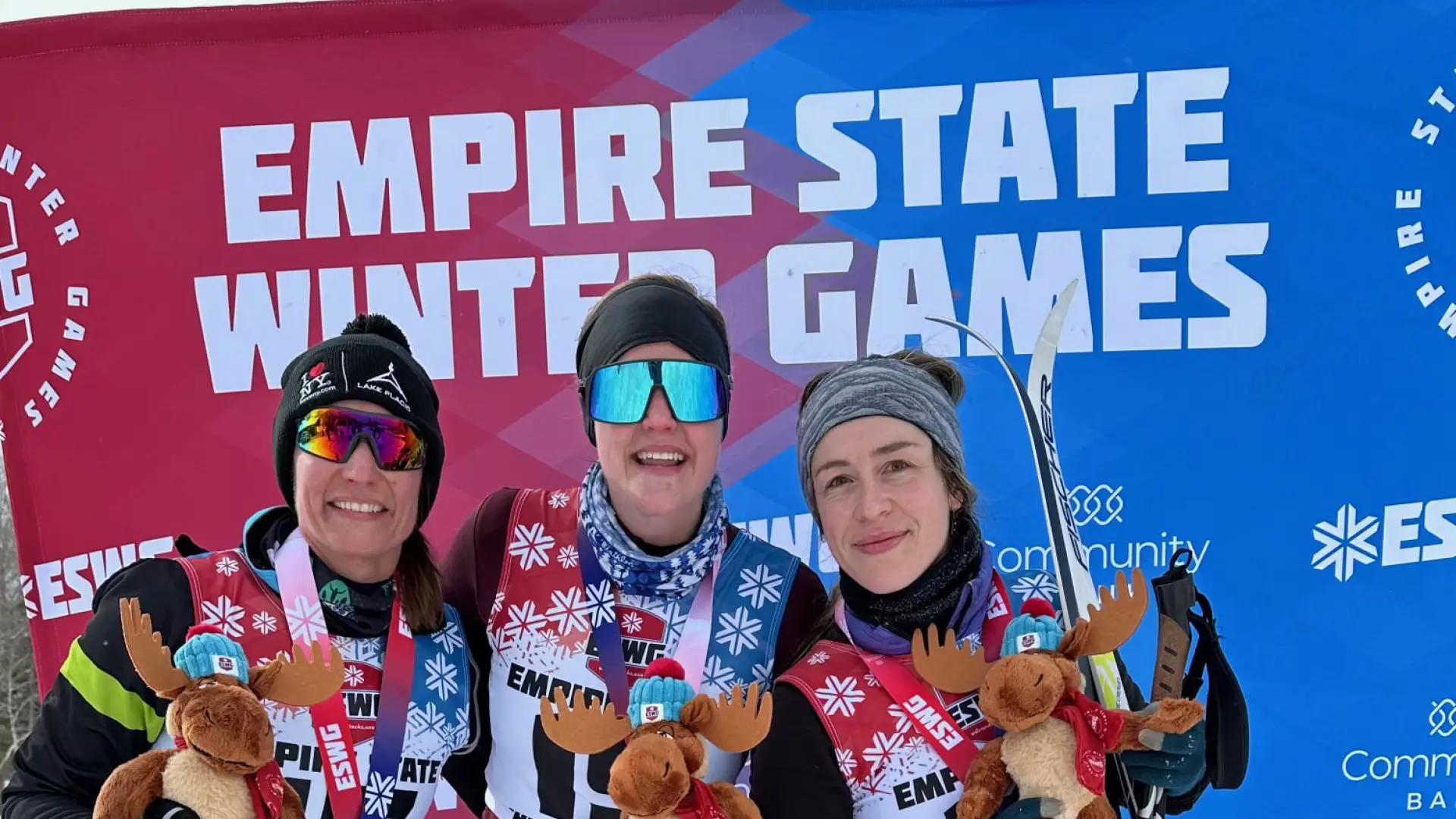 Three skiers proudly pose in front of the "Empire State Winter Games" banner, holding moose plushies and medals, exuding joy and accomplishment in snowy attire.