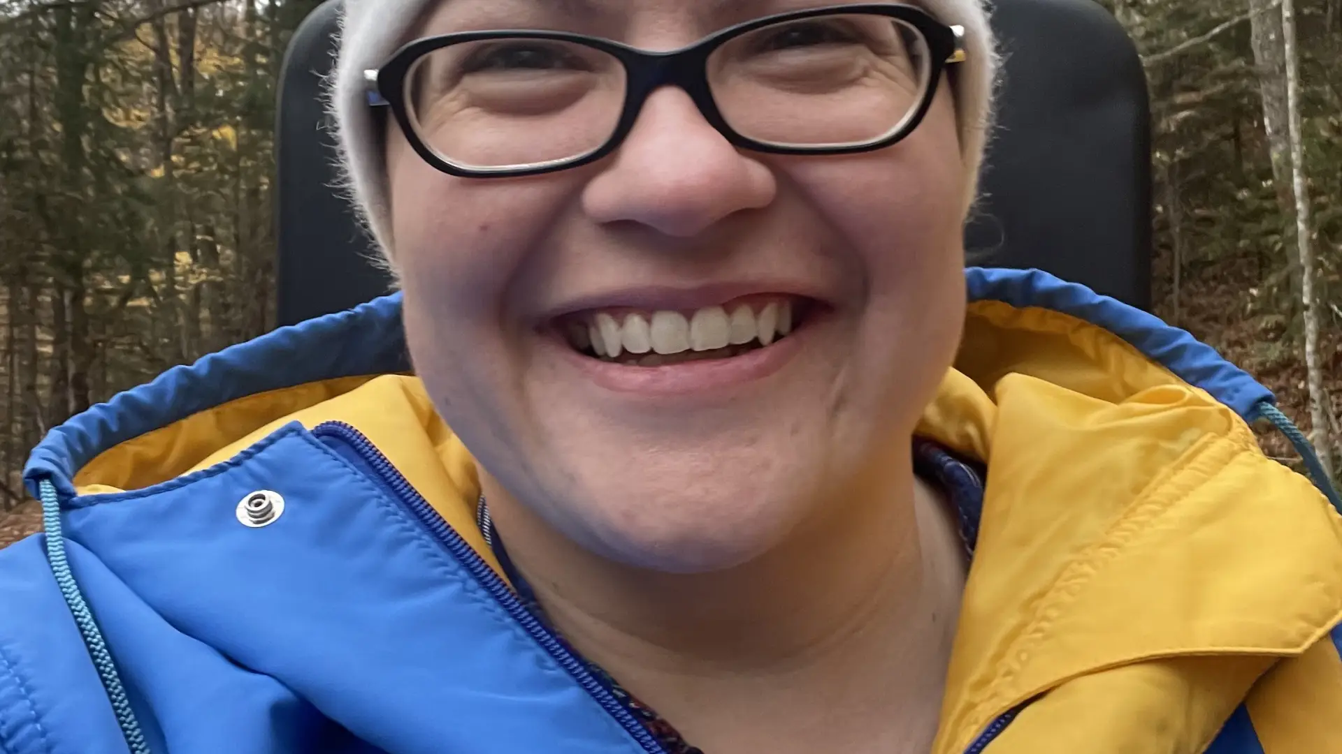 A woman in a bright blue and yellow parka and 1980 Lake Placid Olympic winter hat grins on a ride.