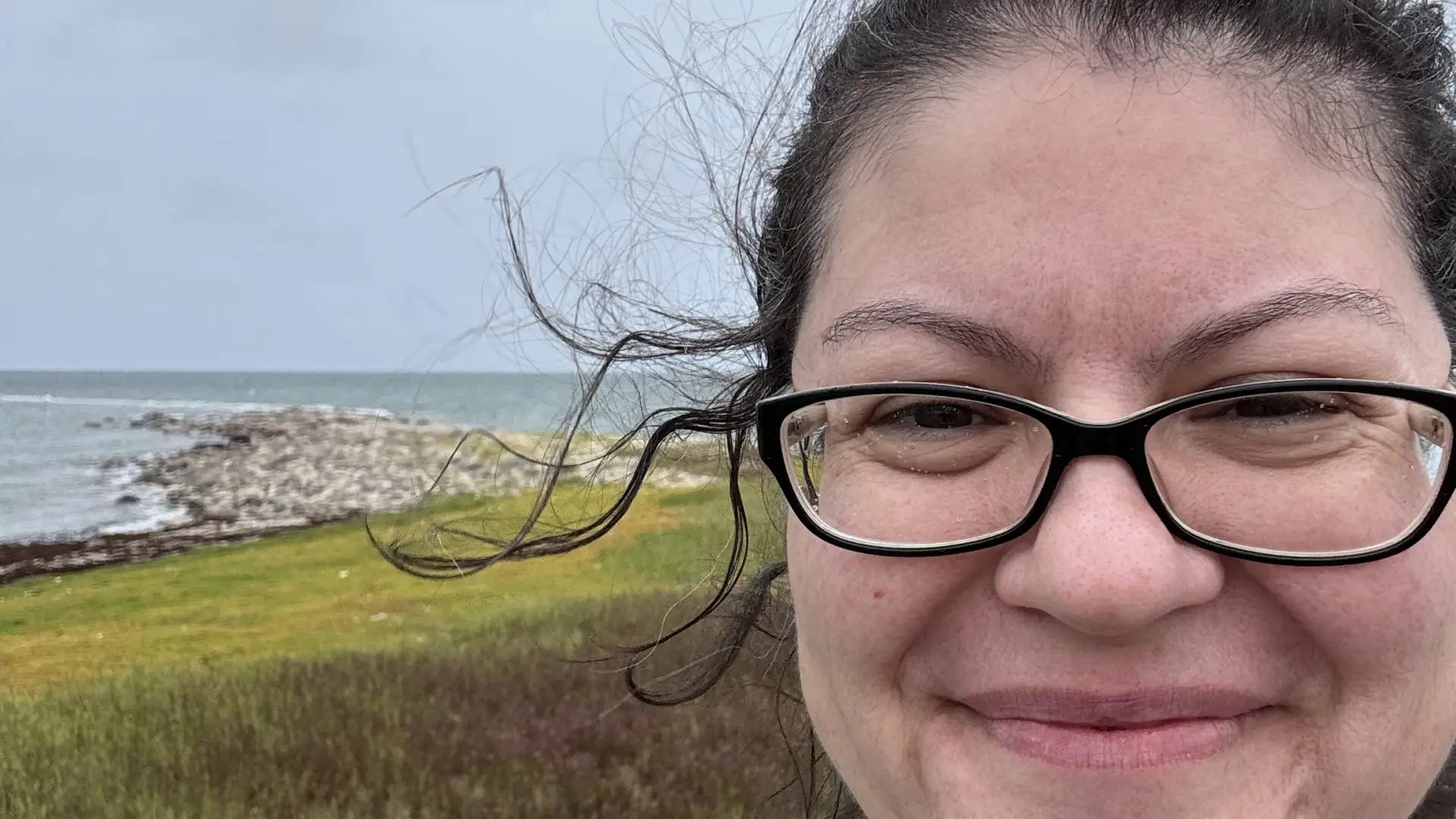 A woman with windblown hair smiles in front of a grey coastline.