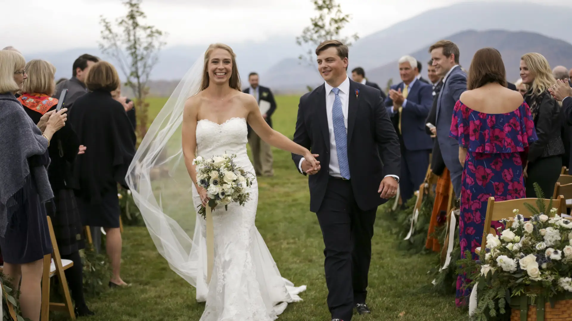 A bride and groom walk away from the alter while holding hands and smiling at friends and family and the mountain backdrop