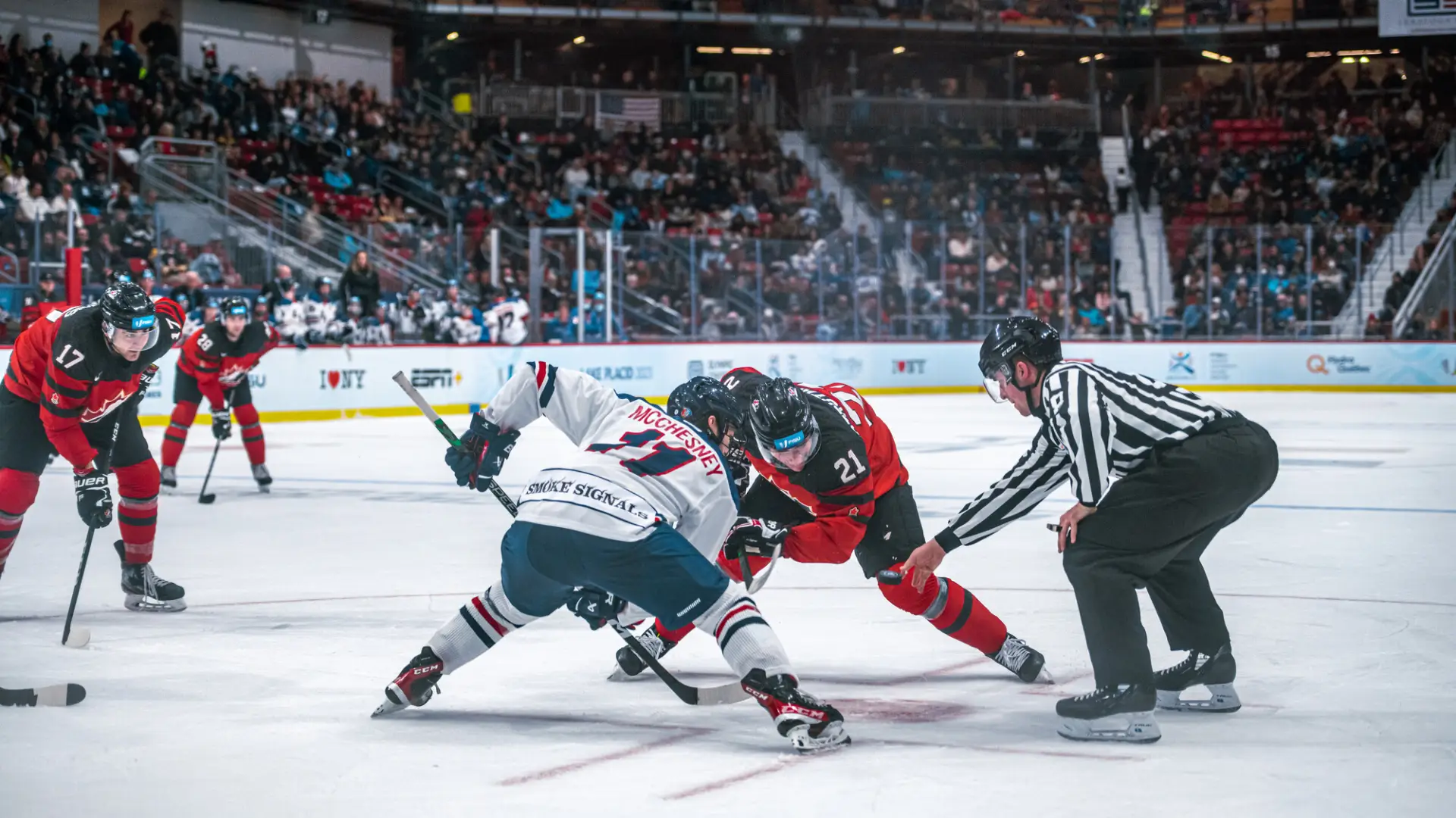 Referee drops puck for two hockey players, one in a white uniform and the other in red, on the ice of skating rink with a packed crowd