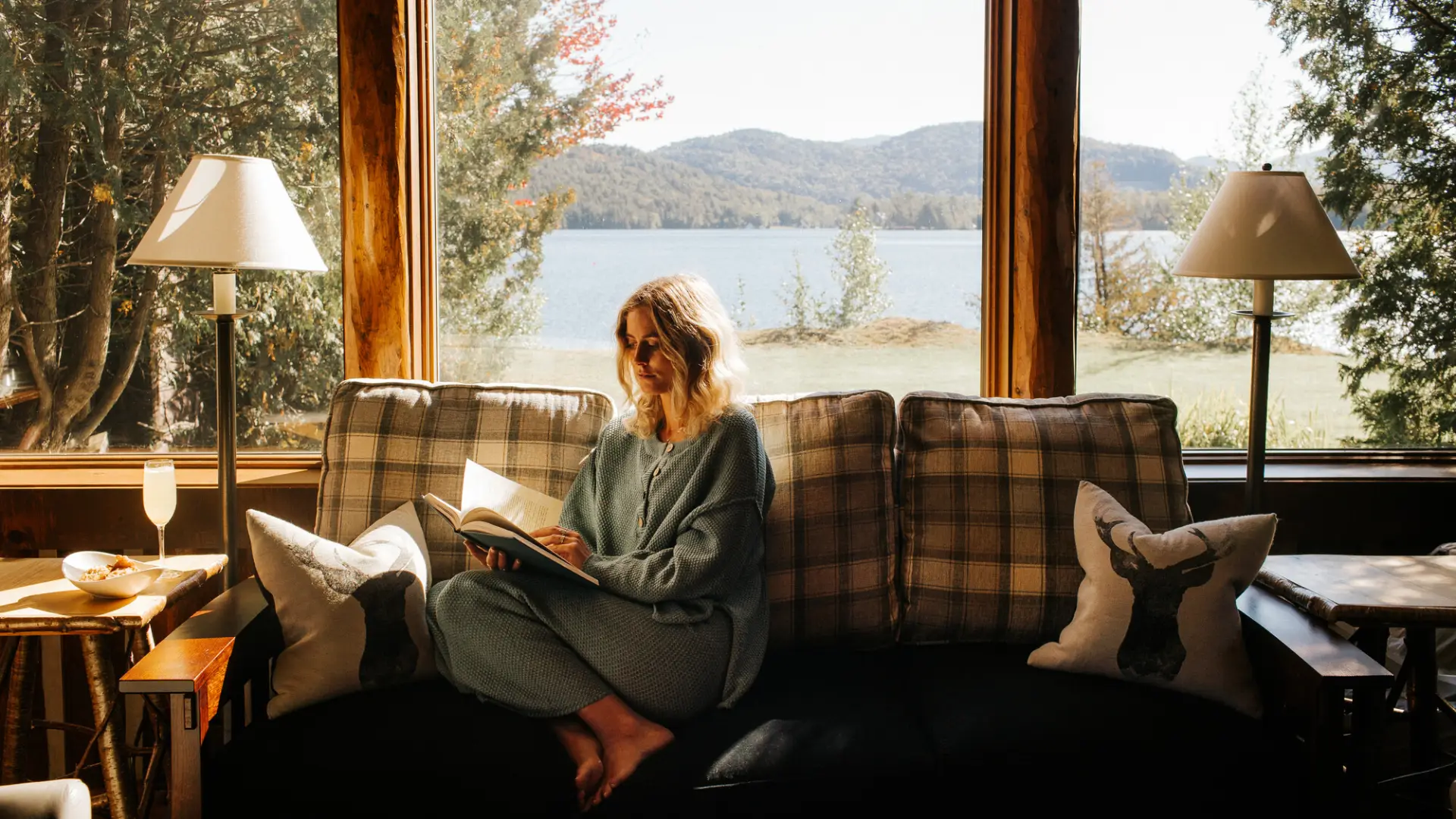 Blonde woman sits on couch with flannel print in loungewear reading a book in front of a window with lake and mountain view through window