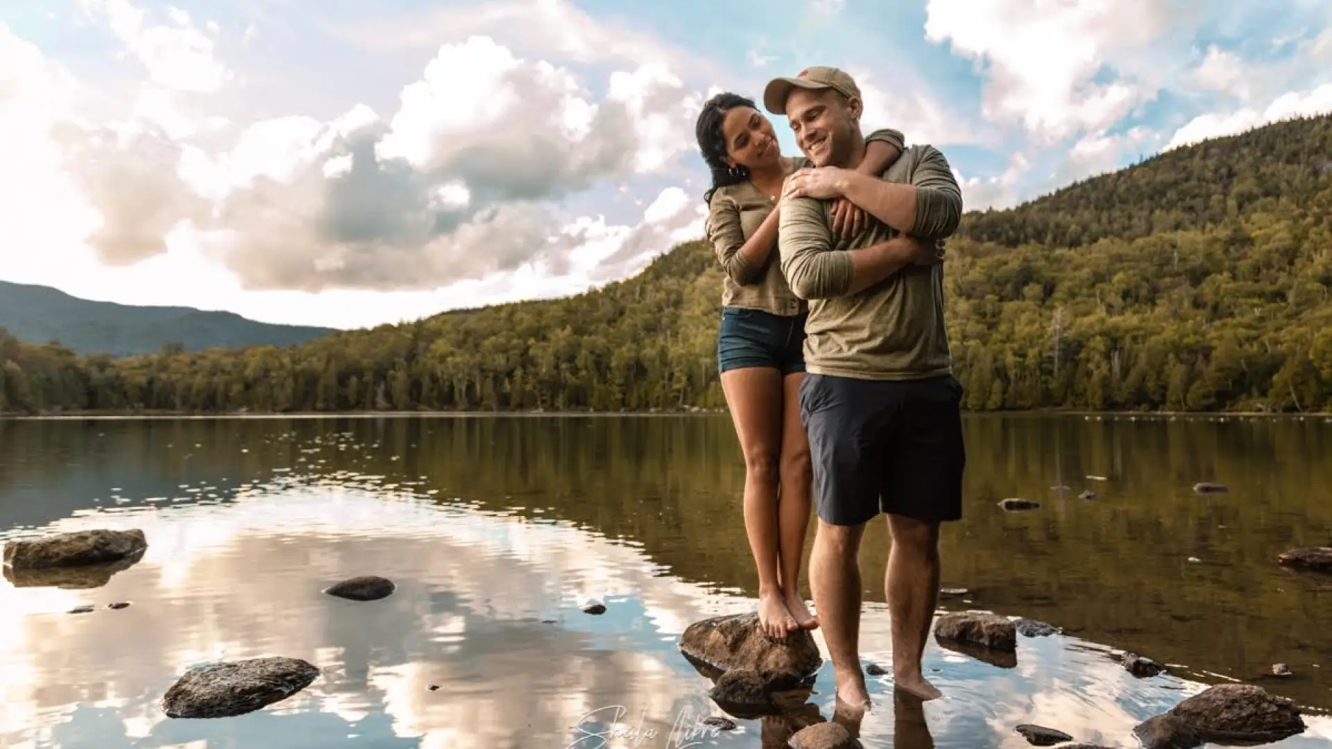 couple-embracing-at-waters-edge-with-mountains-and-blue-skies-in-the-background