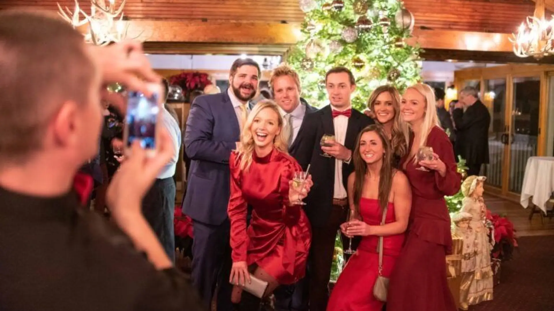 Man takes photo on a cell phone of a group of young people, women in formal dresses and men in suits, gathered together smiling in front of a decorated and lit Christmas tree