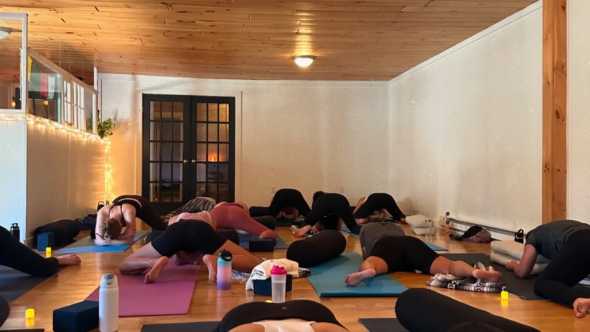 A yoga class in a studio with wood floors and a wood ceiling. Several people are on yoga mats performing a seated forward fold.