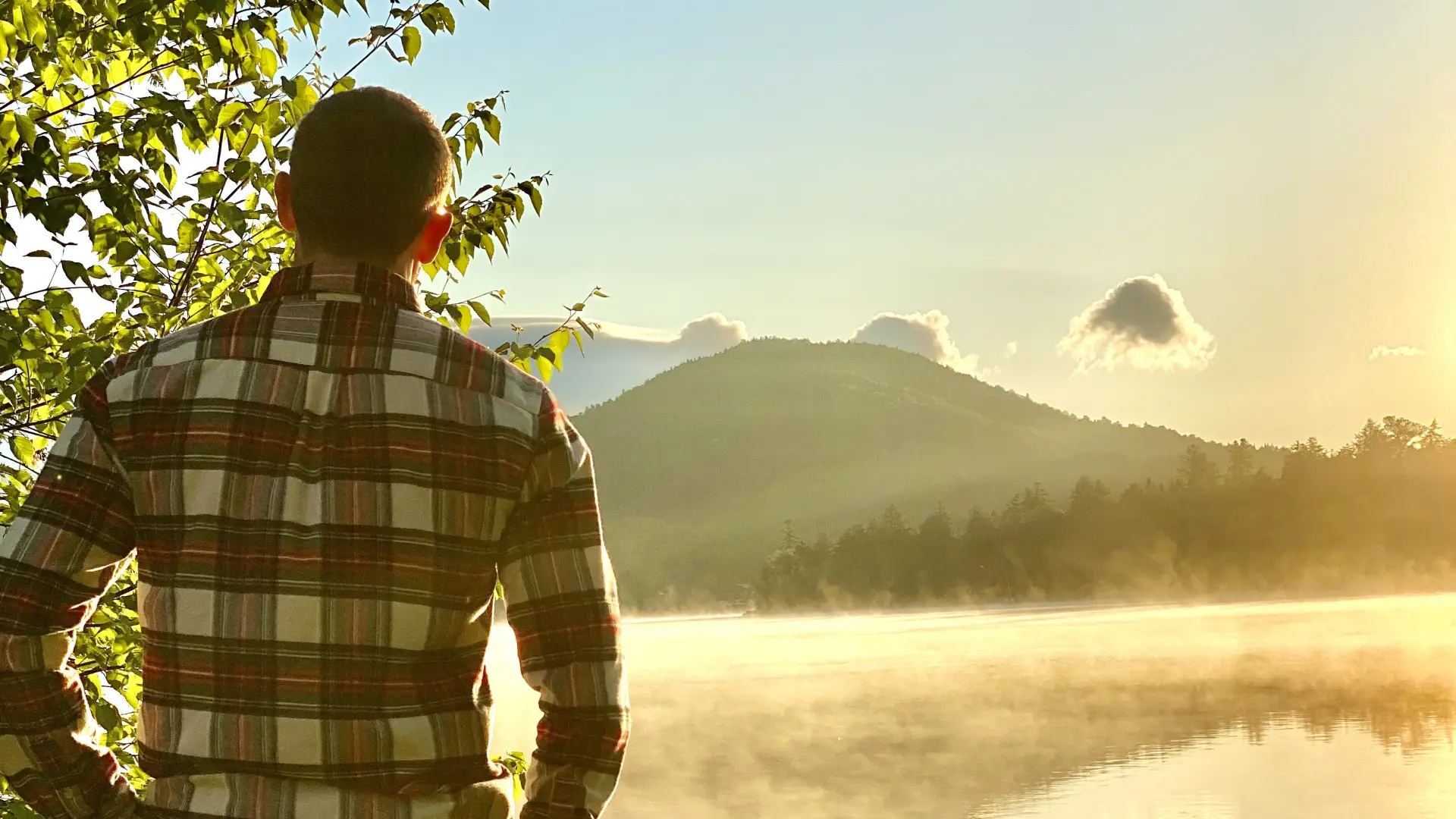 A man in a flannel looks out over a misty lake and mountains.