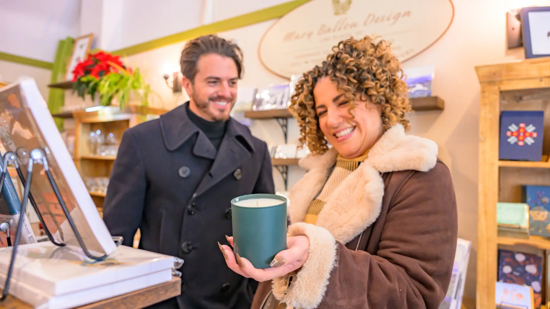 A man and a woman in winter coats smile as the woman holds a candle in a small, quaint gift shop.