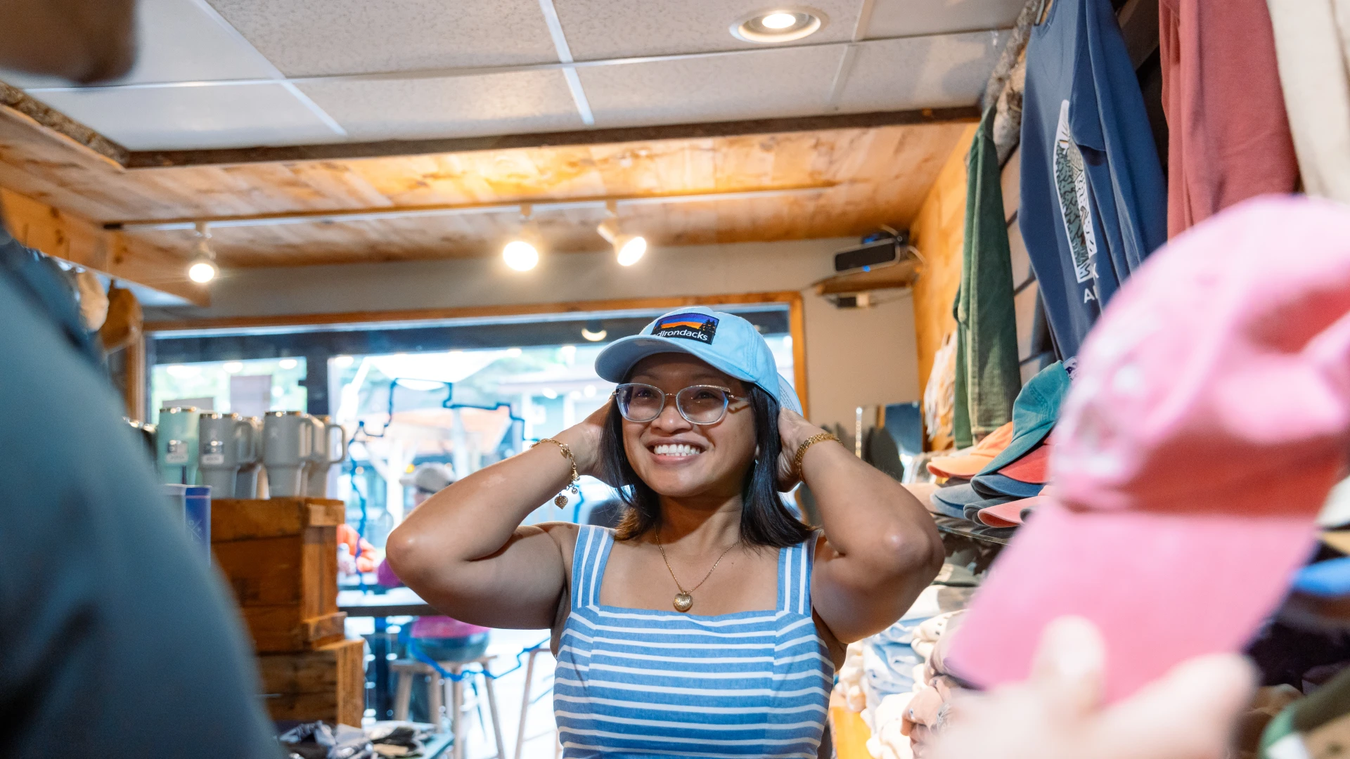 A smiling woman wearing a blue-and-white striped dress and glasses tries on a light blue baseball cap inside a shop.