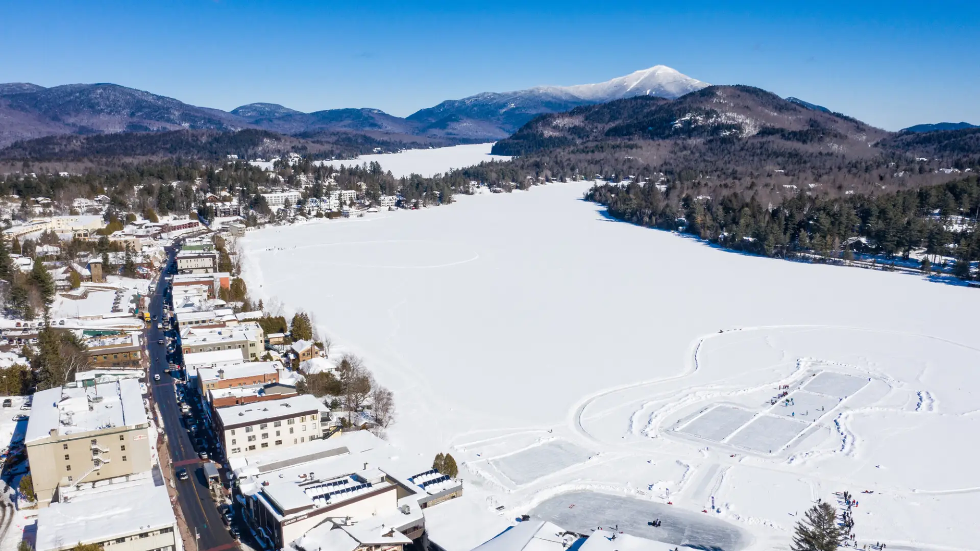 A frozen lake surrounded by a mountain town and mountains in the background on a clear day. 