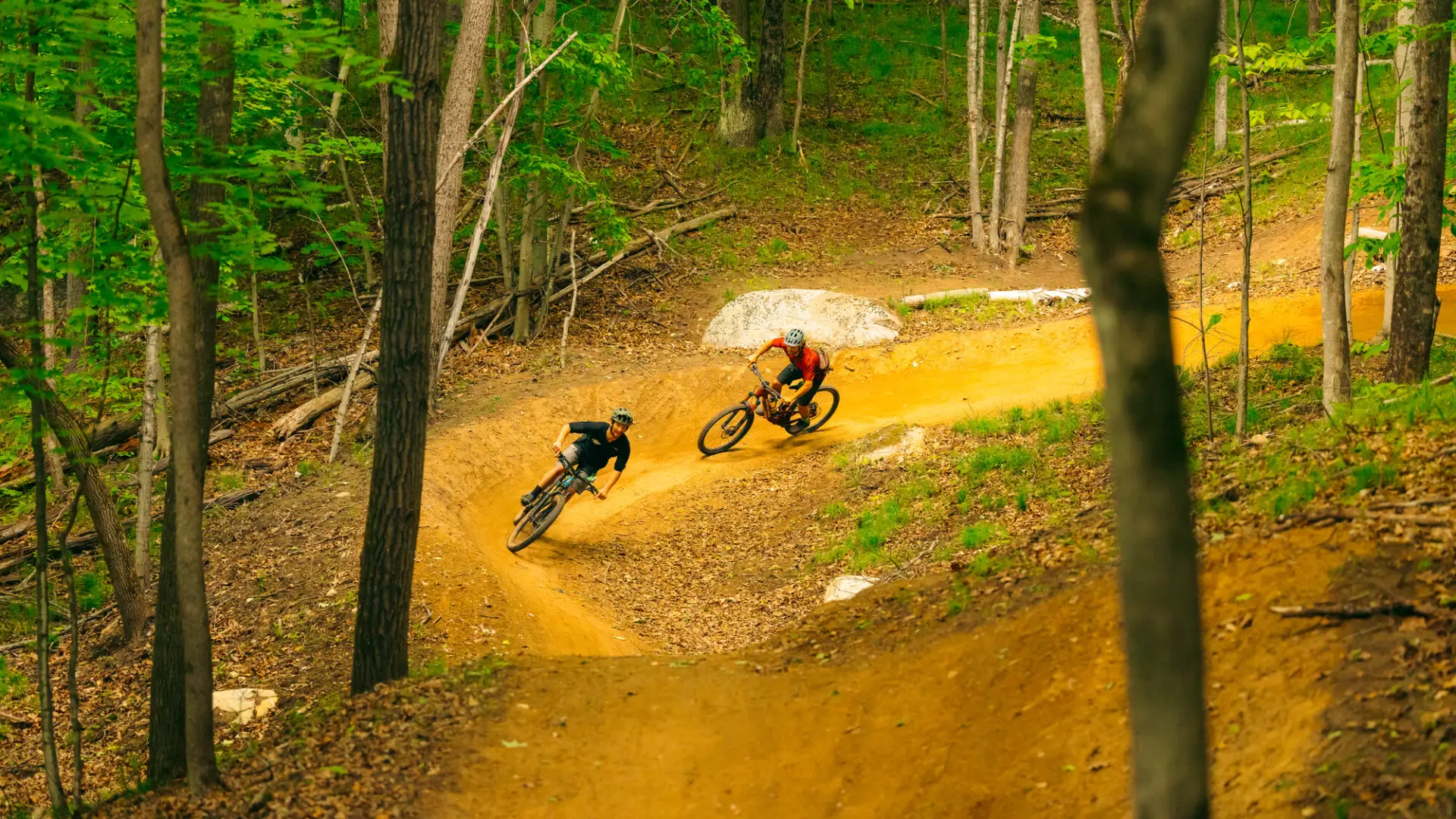 Two mountain bikers ride single file around berm on dirt track in woods