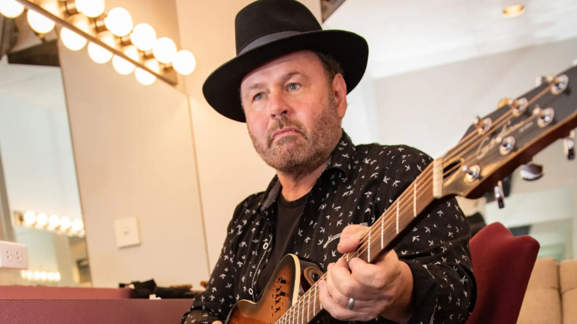 A middle-aged man with a beard wearing a black hat and a black shirt with white birds on it plays an acoustic guitar.