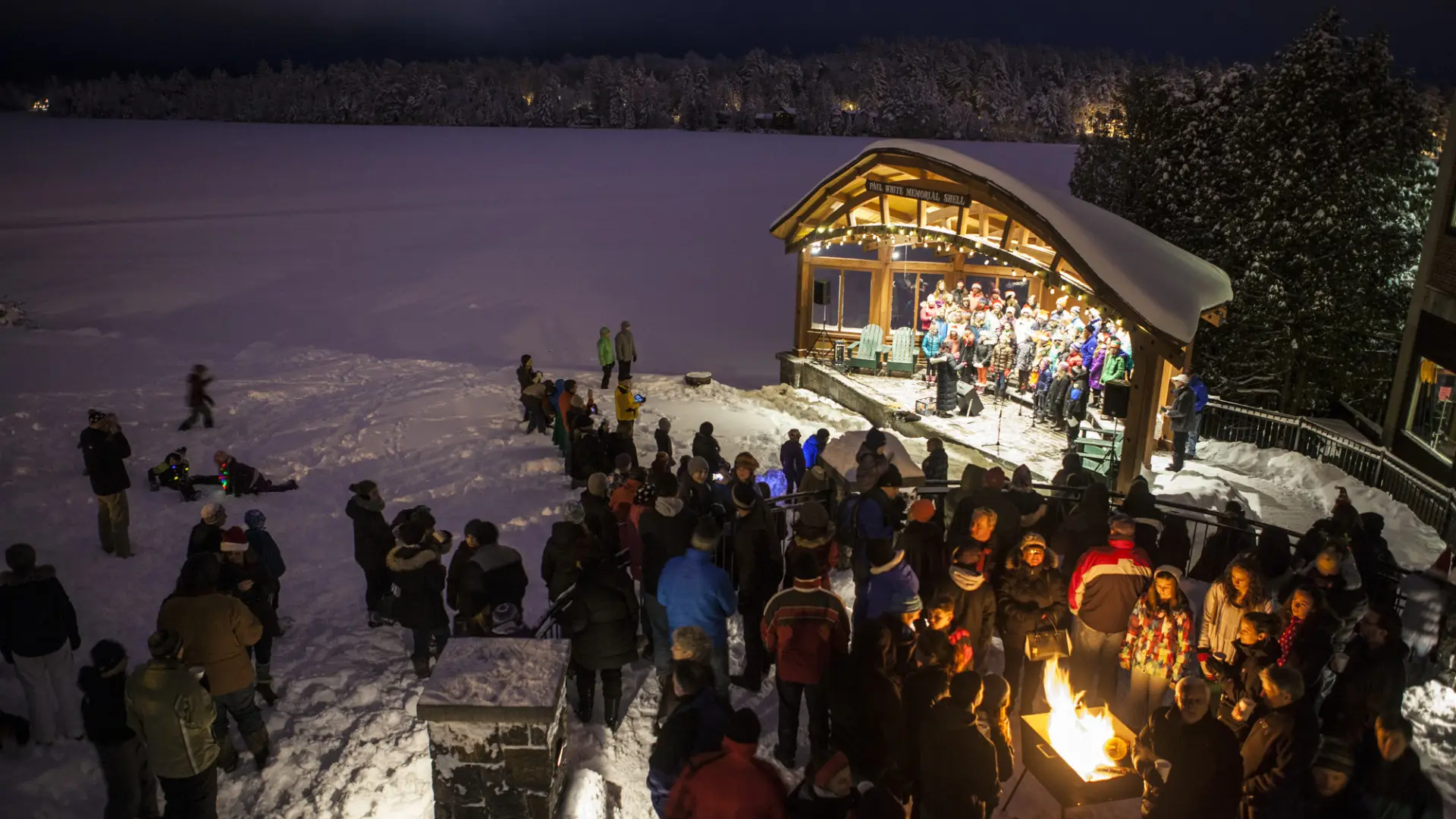 A crowd watches christmas carolers in a band shell next to a frozen lake.