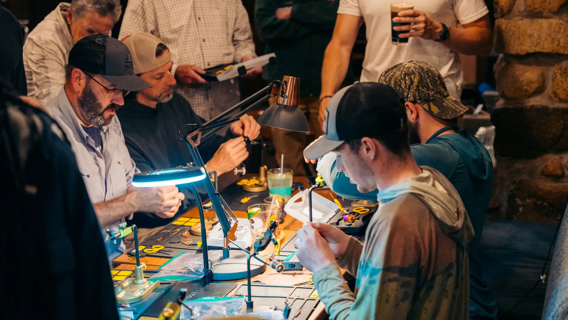 Group of people work on tying fishing flies under small lamps around a table at the Ausable River Two-Fly event