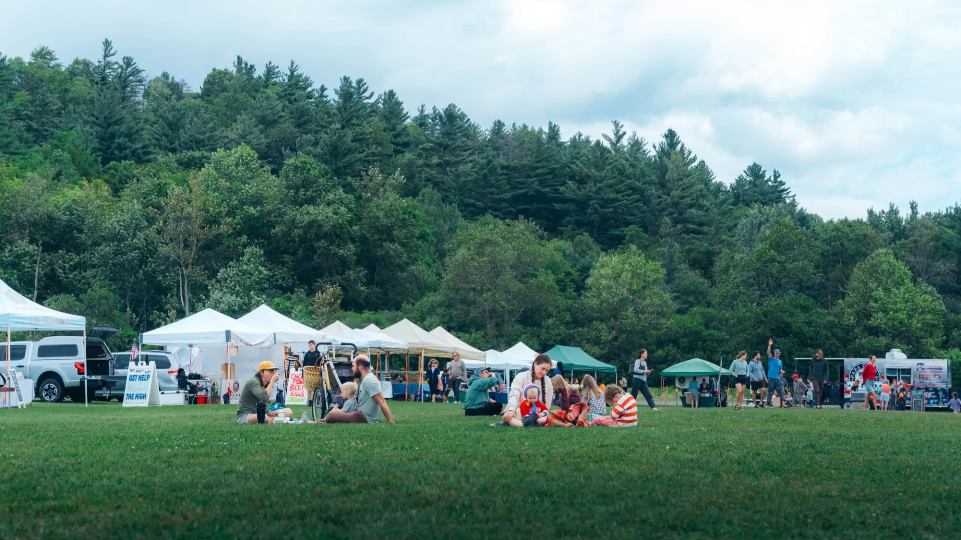 A wide shot of people enjoying a grassy field at an outdoor market with white vendor tents and a backdrop of pine trees.