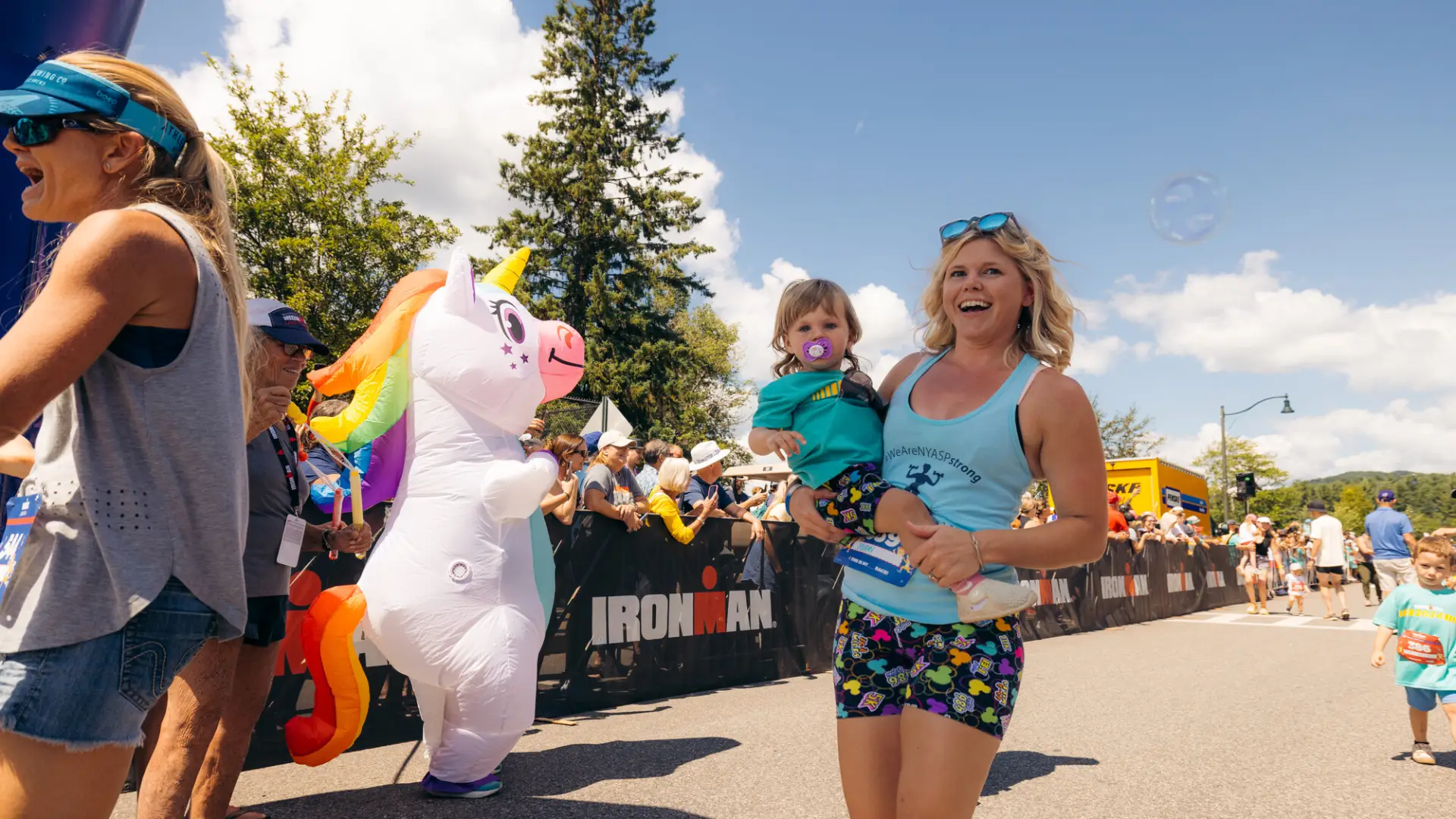 Woman in tank top and shorts holding baby walks past person in unicorn costume at race