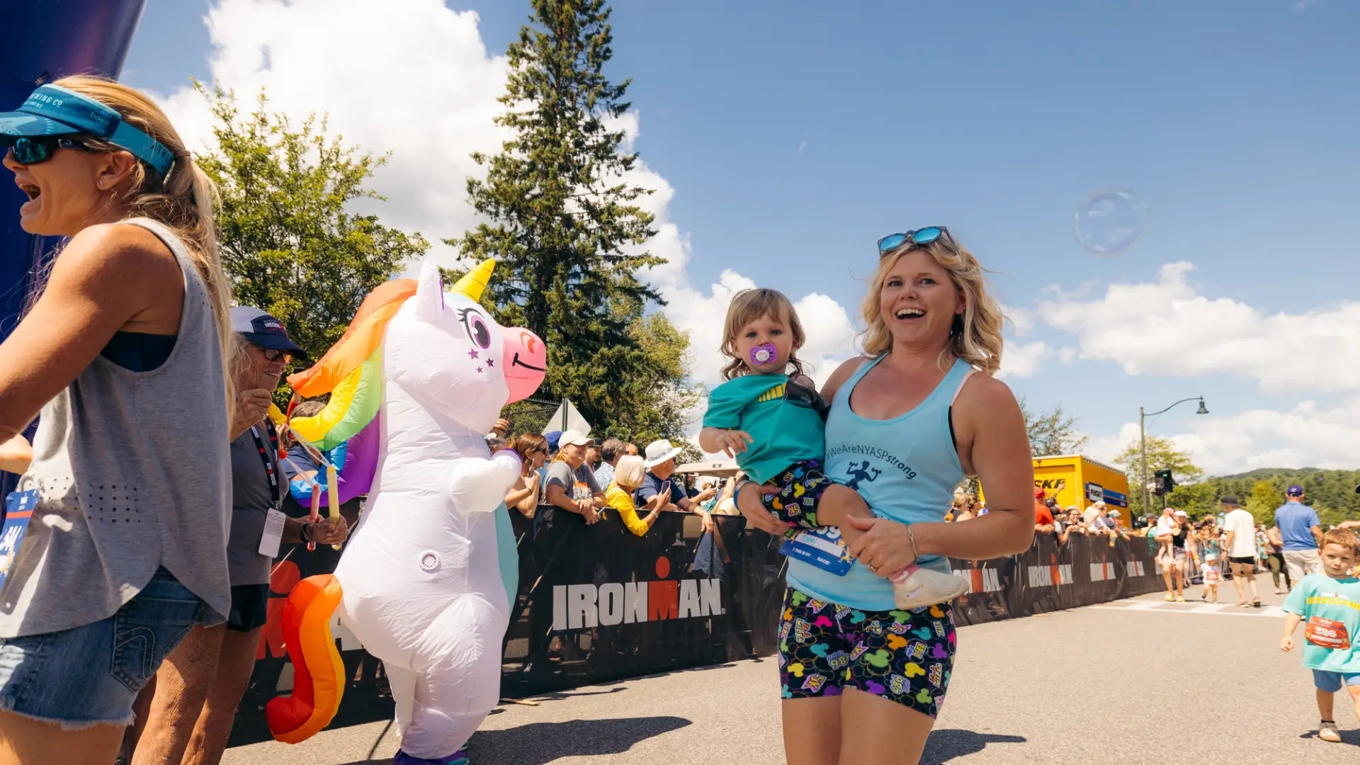 Woman in tank top and colorful shorts holds toddler while walking across finish line with inflatable unicorn behind