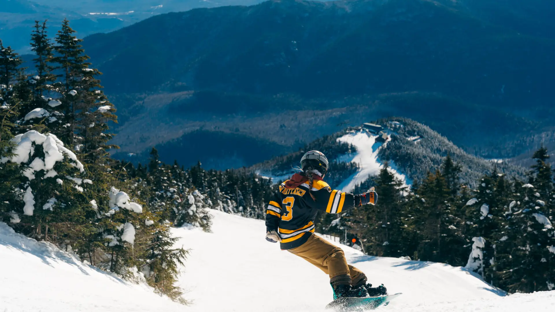 A woman in a bruins jersey snowboards down a mountain.