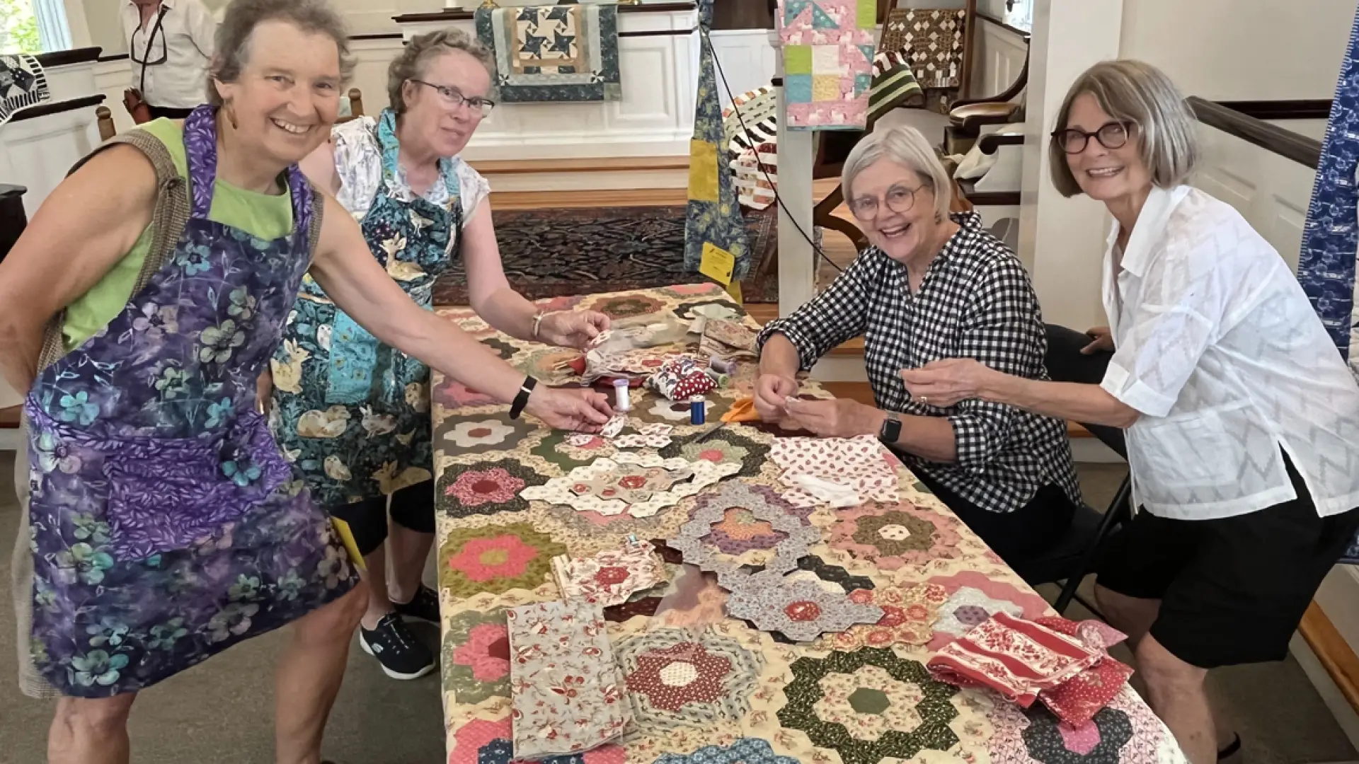 Four senior woman gathered around a table covered in a quilt and quilting materials look at camera smiling