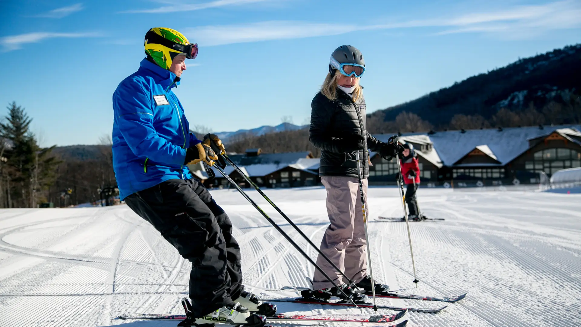 Ski instructor on skis in blue coat shows new female skier on skis how to lean back in your boots