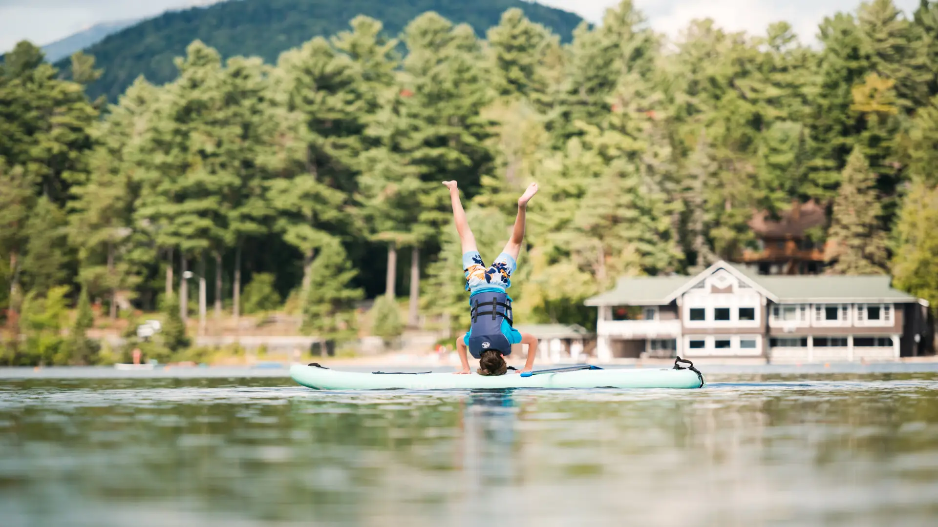 A teenager in swim clothing and a life jacket does a handstand on a stand-up paddleboard on a lake.