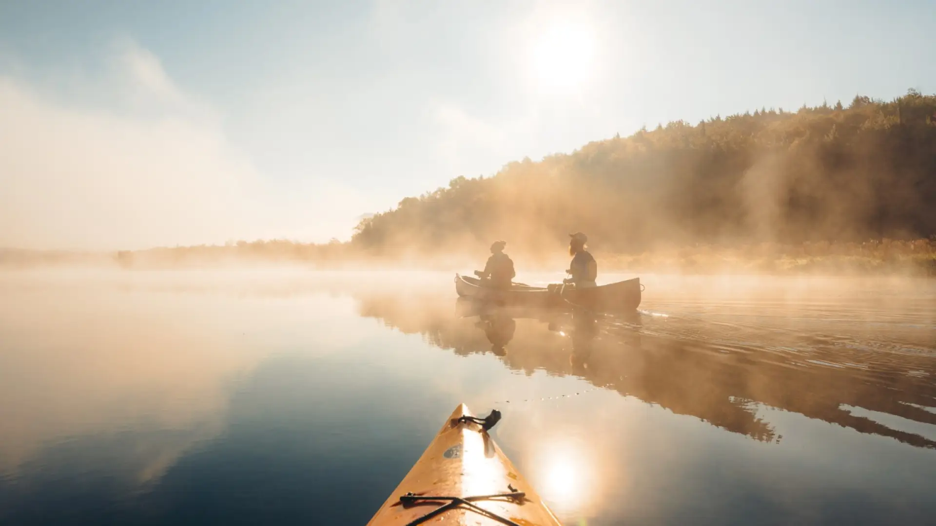 Two people paddle canoe on mirror-like water with fog seen past bow of orange kayak
