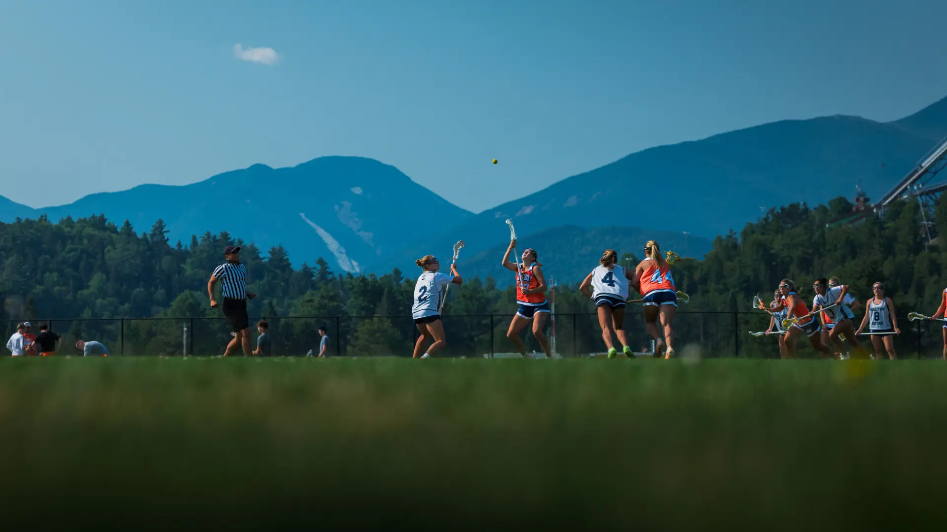 Two women holding lacrosse sticks look up at yellow lacrosse ball in the air during play on field with mountain views