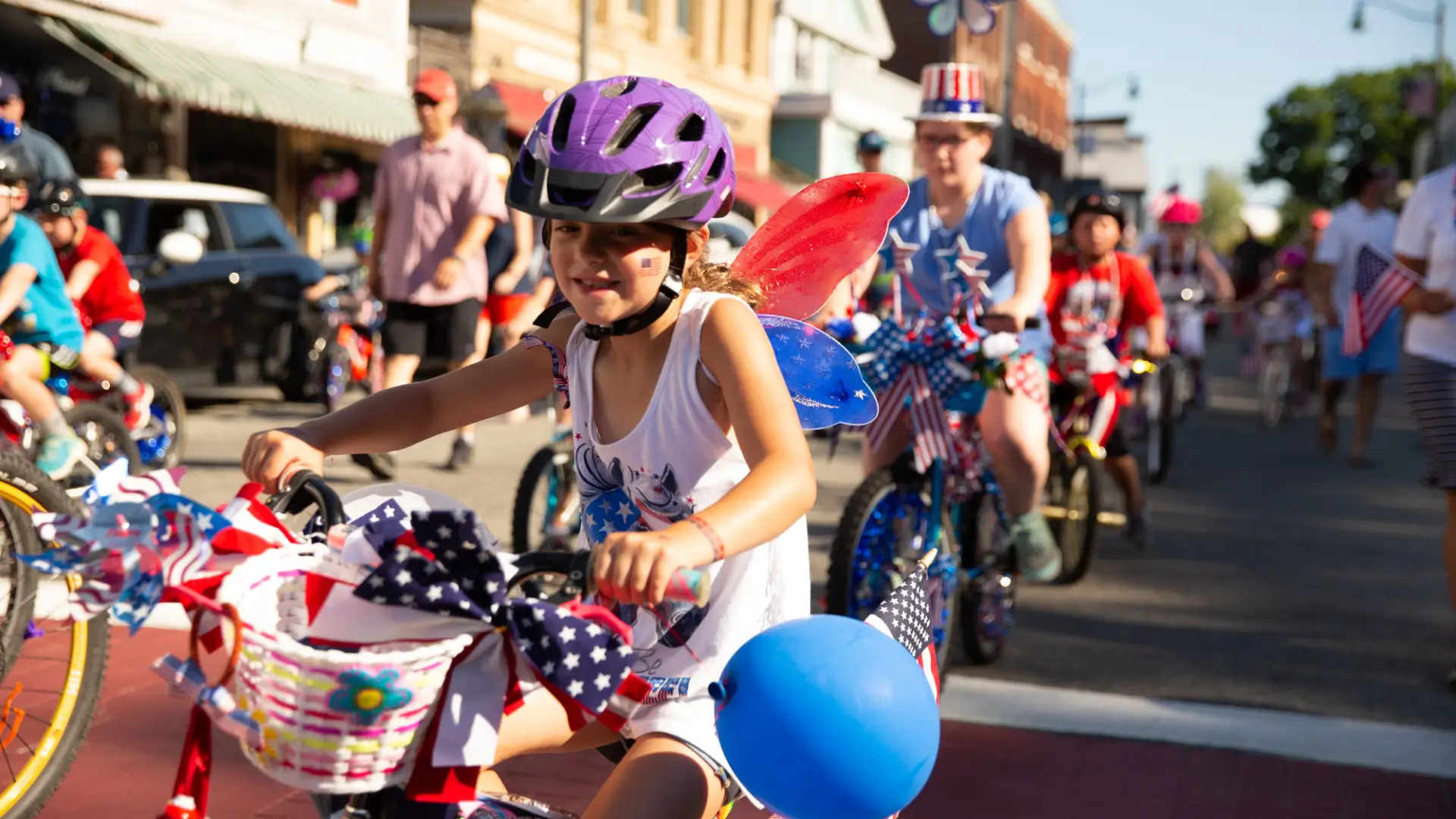 A young girl decorated in July 4th colors and ribbons rides a bike in a parade.
