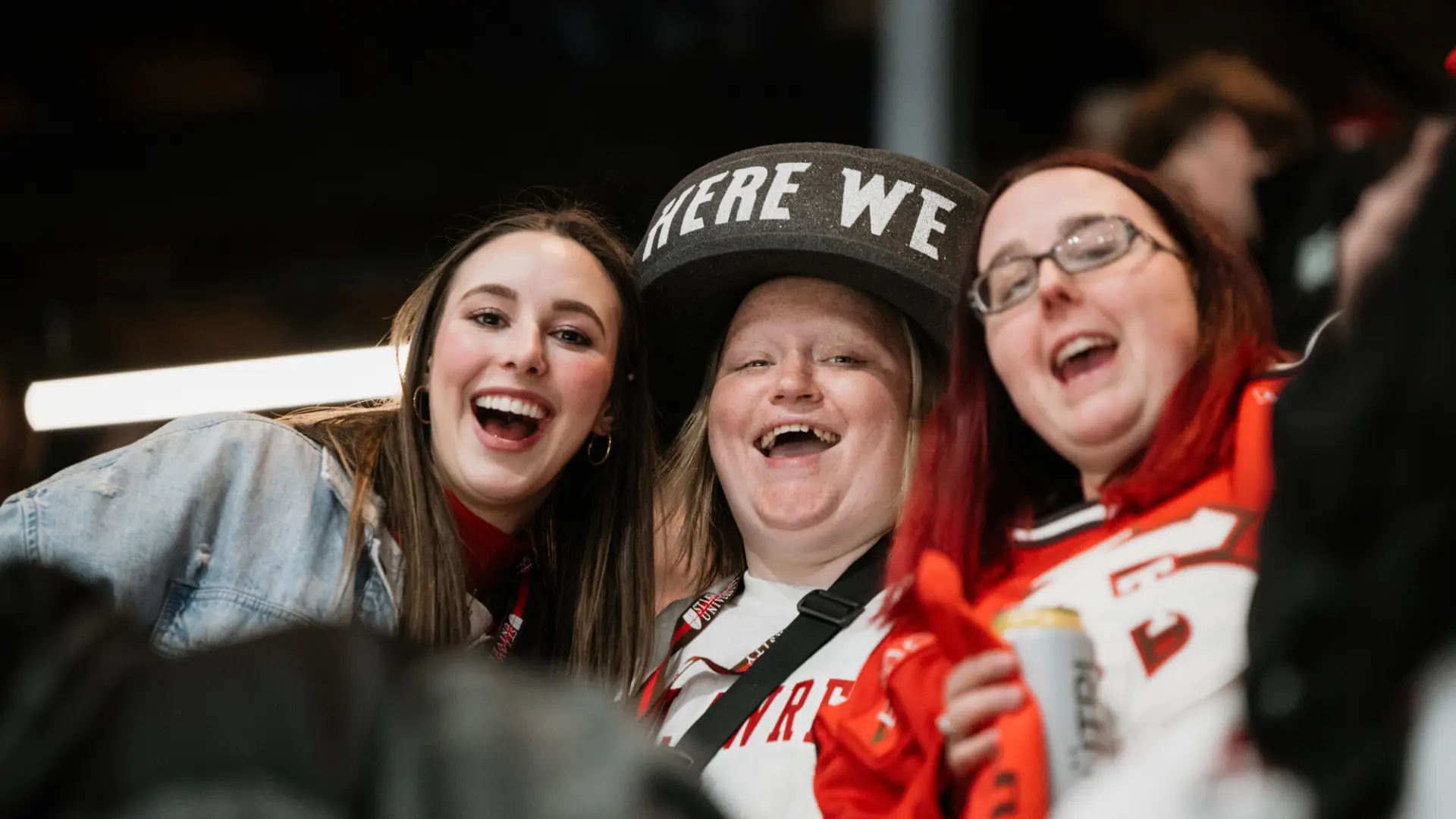 Three young woman stand closely together cheering in the stands of a hockey arena. The woman are wearing hockey jerseys and accessories. One is wearing a black foam hat with white lettering