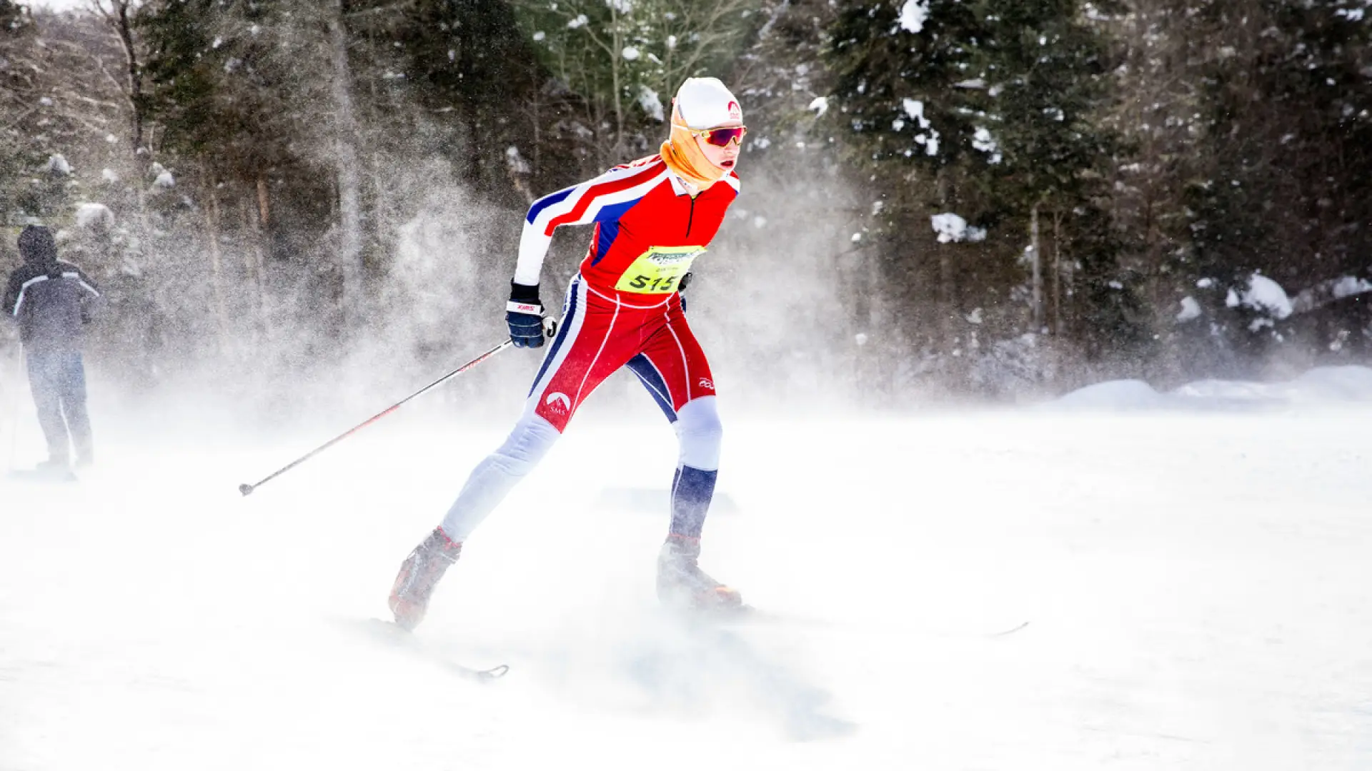 Nordic skier in red race outfit skis through lose snow, kicking up a cloud of snow around them. They wear a yellow race bib on their chest