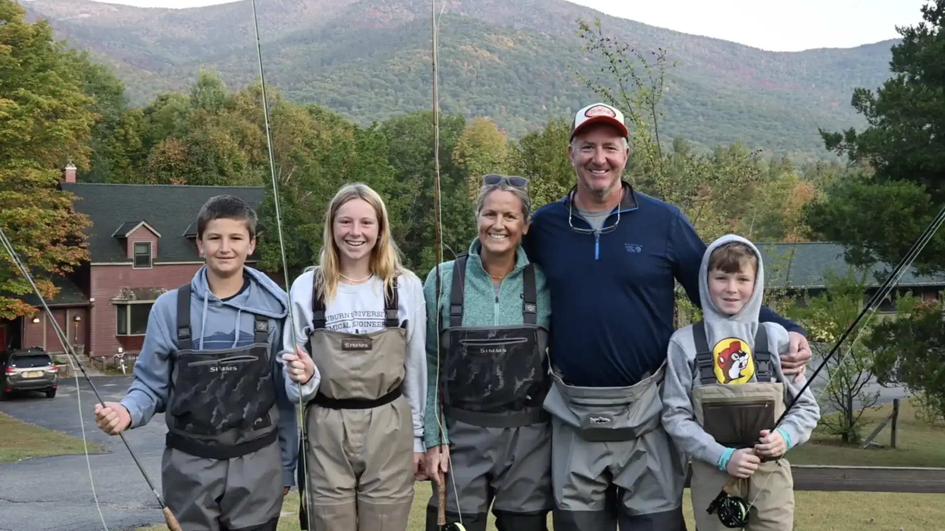A family of five stands outdoors in waders and holds fishing rods, with mountains in the background.