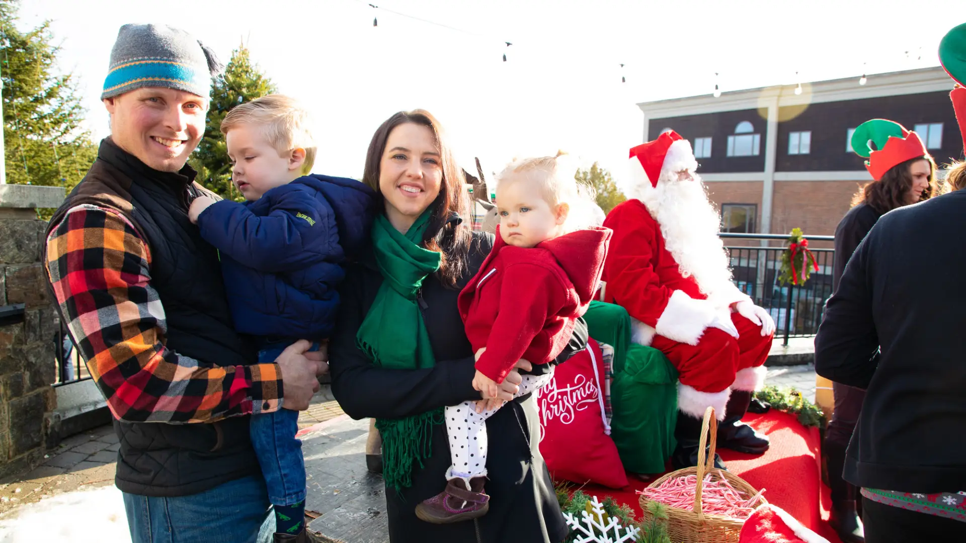 Parents with holding to small children stand outdoors near a Santa Claus seated behind them