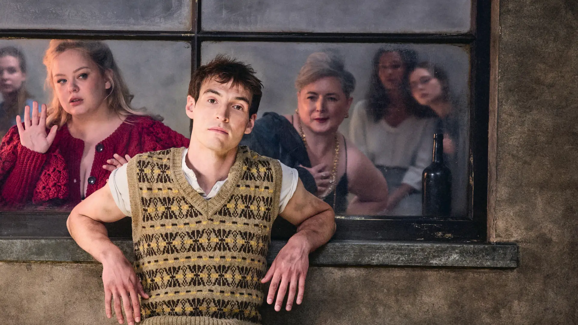 Young man in sweater vest and short sleeves leans against window sill with four women looking out the window behind him