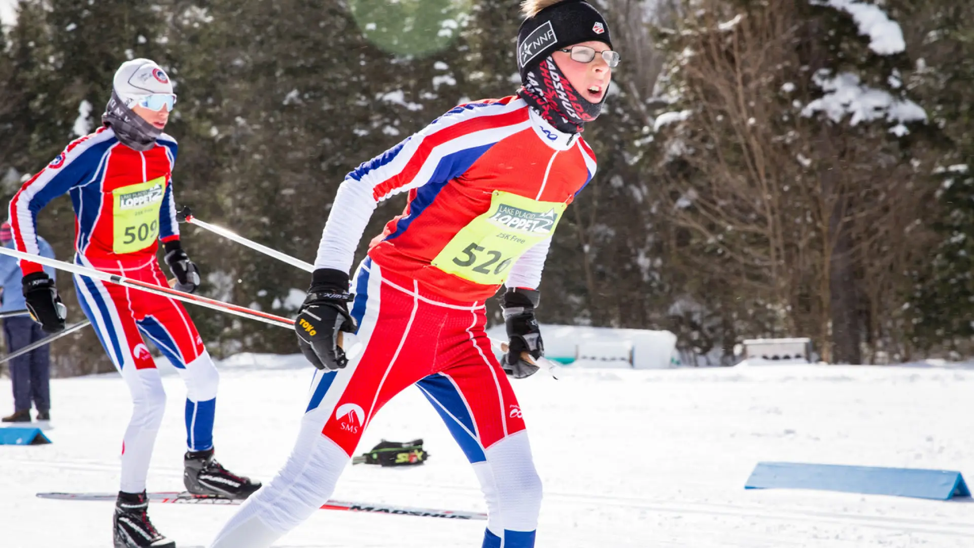 A nordic skier in red, white, and blue cross-country ski gear and a yellow race bib skis past the camera from left to right on a side snowy trail in the woods.