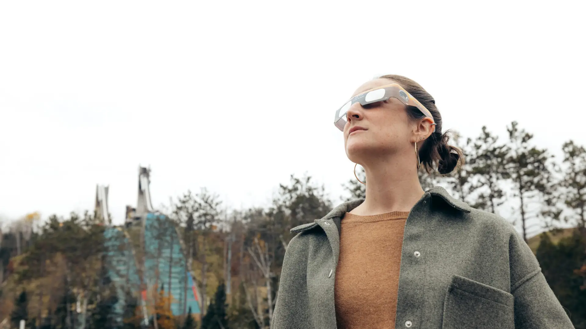 A woman wearing eclipse glasses gazes up at the sky with ski jumps in the background.