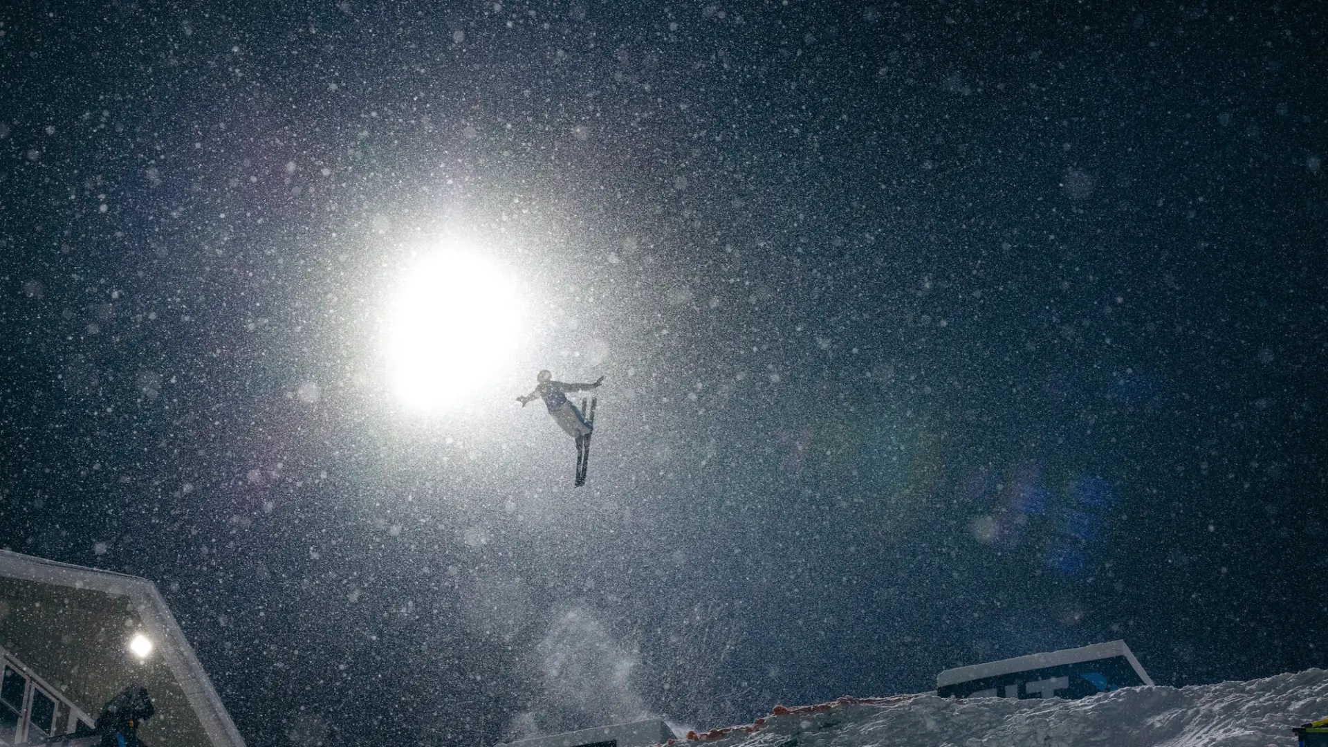 Freestyle aerial skier performs a flip in the air on a dark night with a bright florescent light lighting the skier, ski hill, and snowflakes in the air