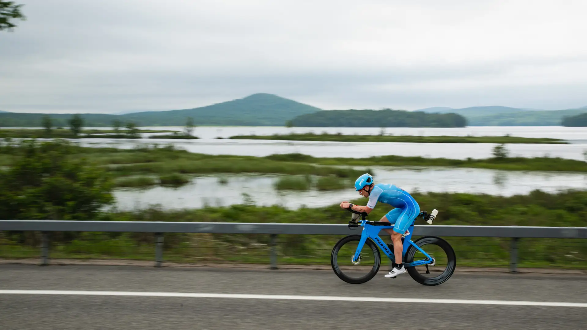 A triathlete in a bright blue speed suit and matching helmet rides a blue aerodynamic bike along a lakeside road.