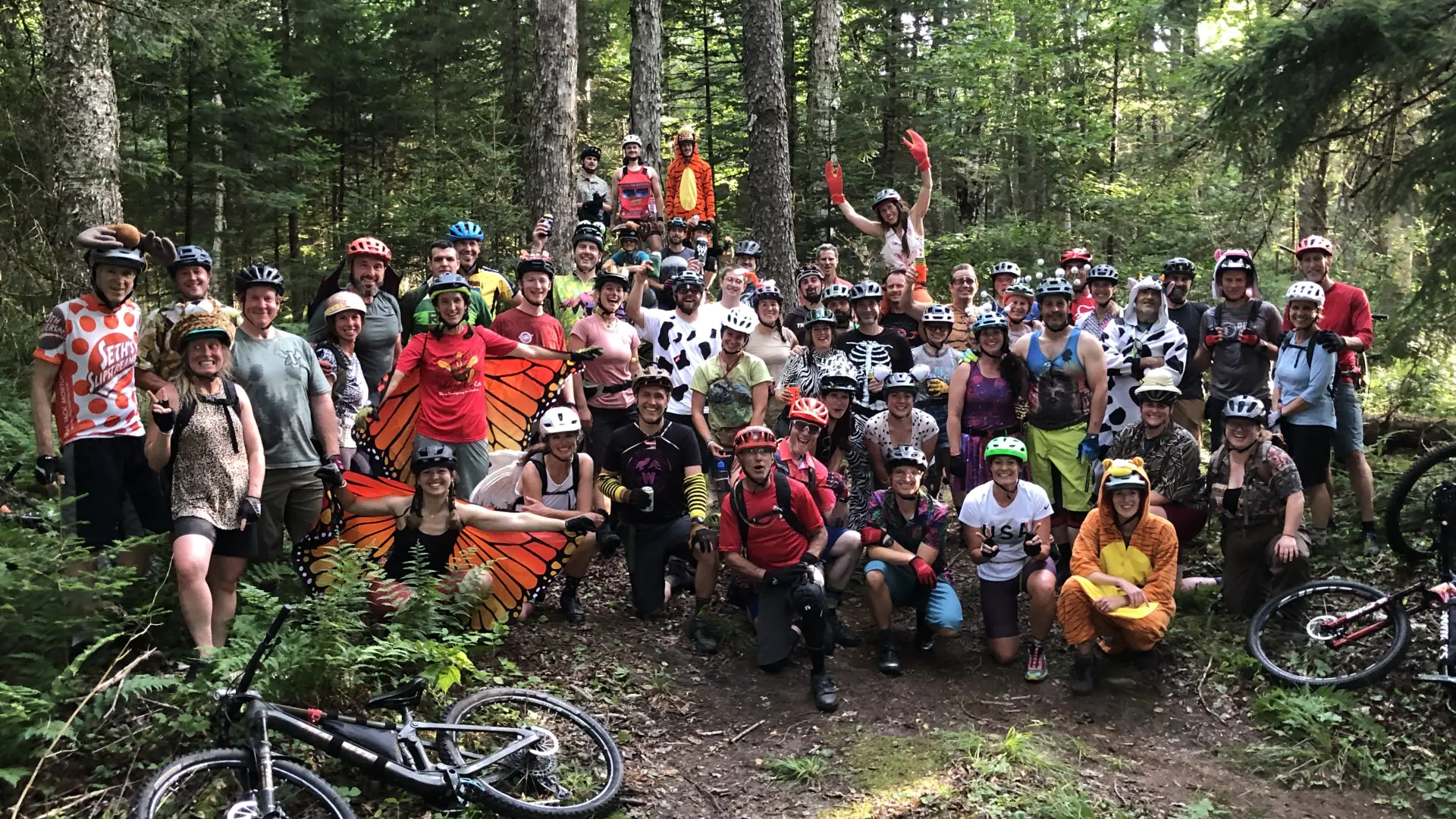 Large group of people, many dressed in a combo of animal costumes and biking gear, pose for photo in the woods with bikes