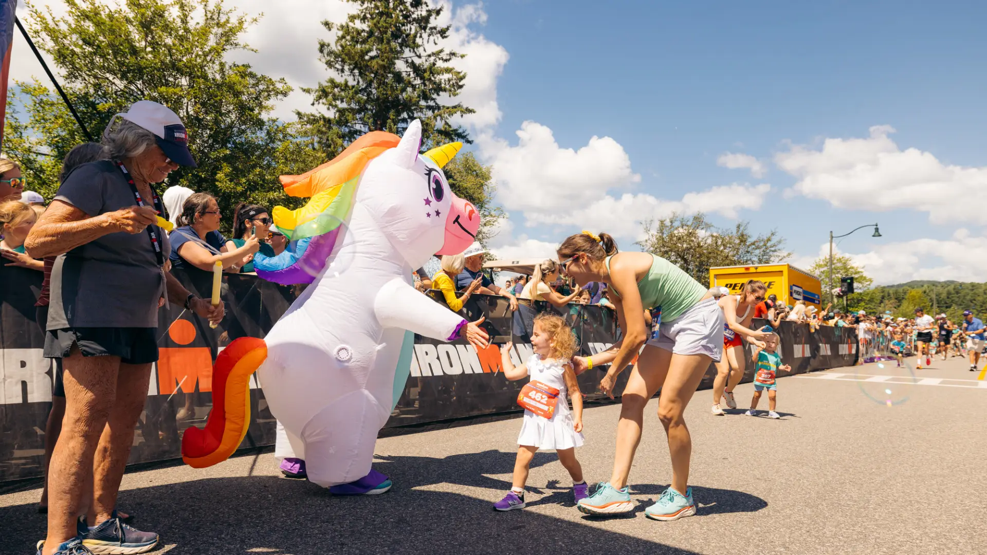 Young girl in white dress accompanied by woman in tshirt and shorts gives a high five to a unicorn mascot while race volunteer waits with finisher medal