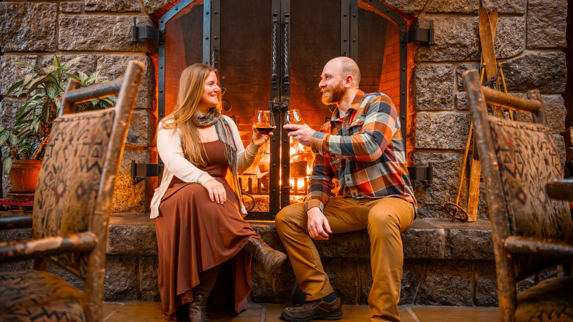Couple sits in front of stone fireplace with raised glasses of red wine