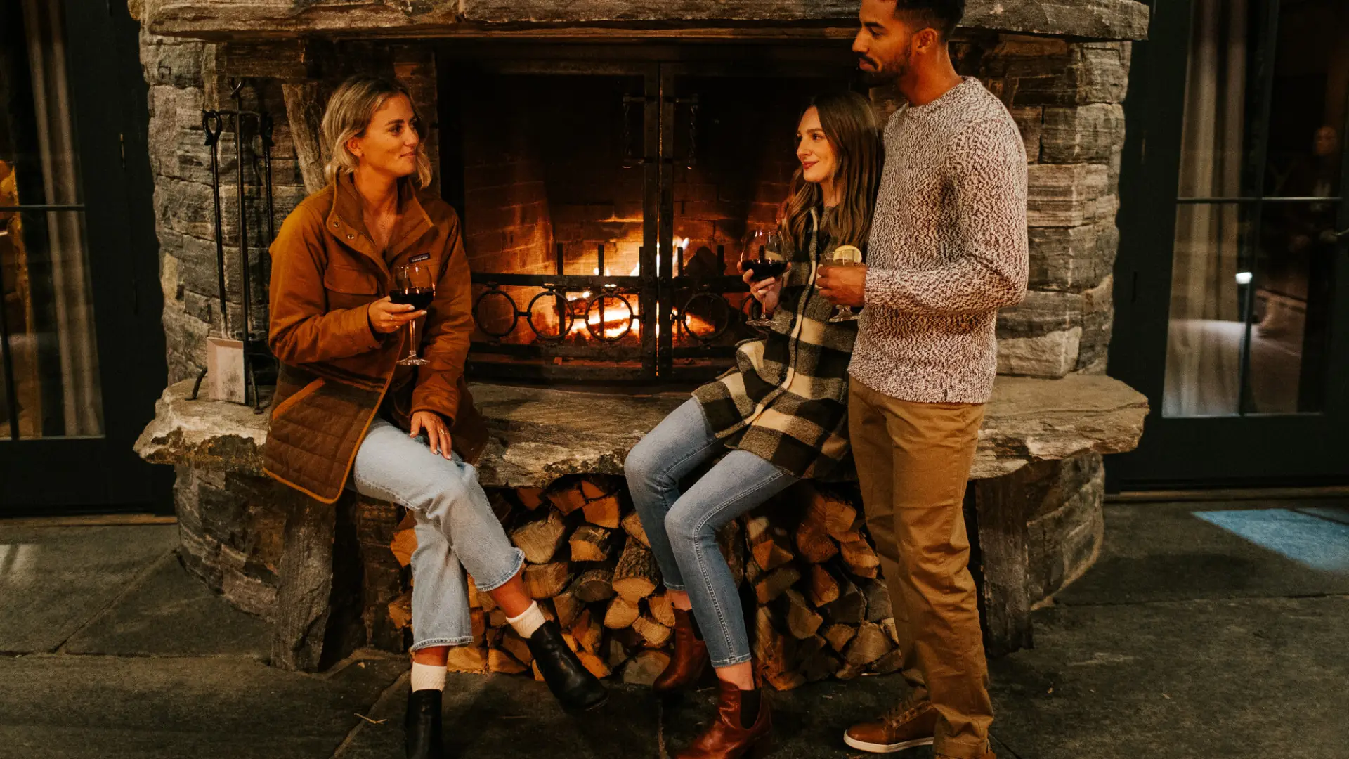 Three friends share drinks at a fireplace.