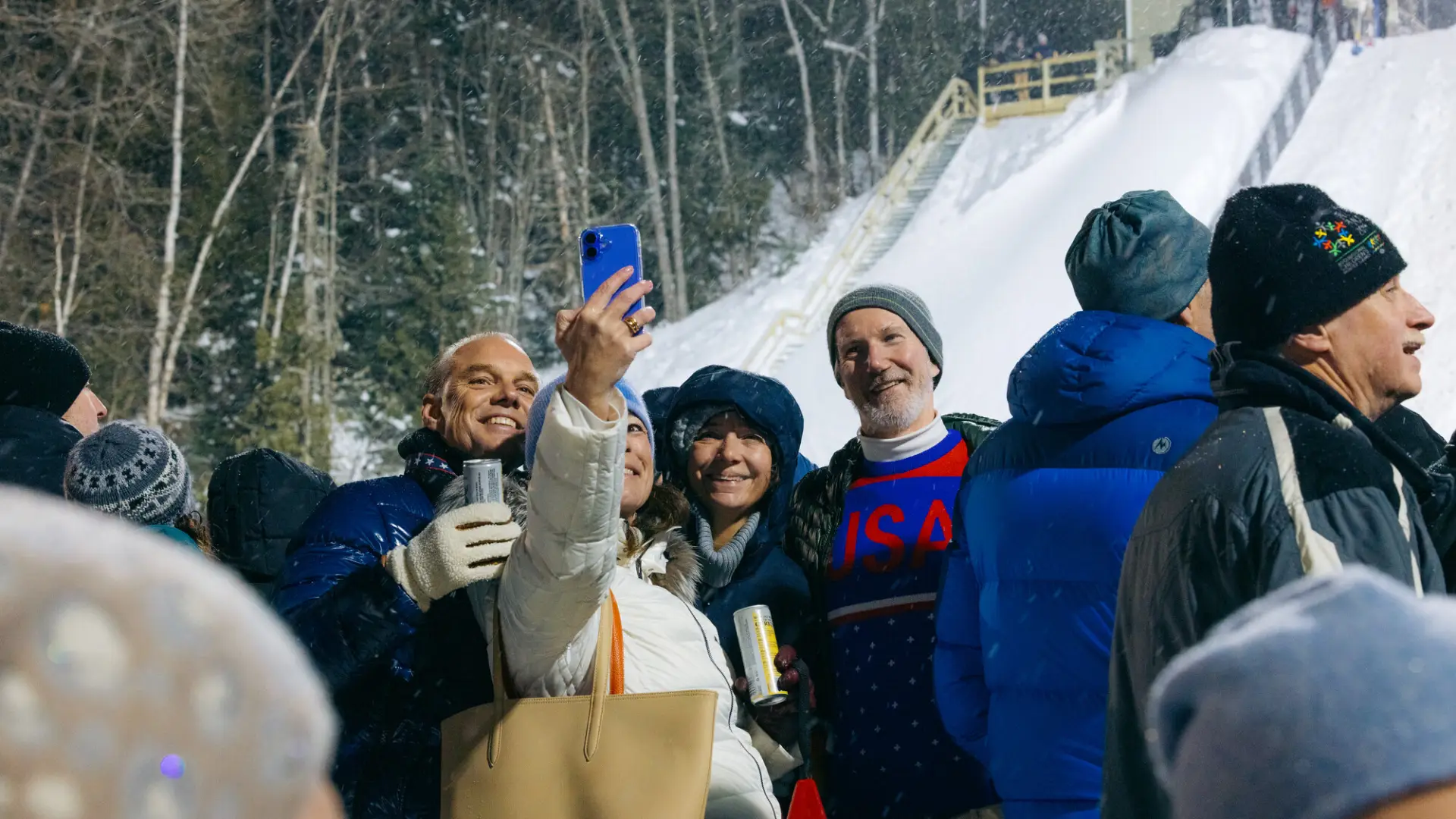 Group of people in winter outwear stand outdoors in snow among a crowd taking a picture at a nighttime event