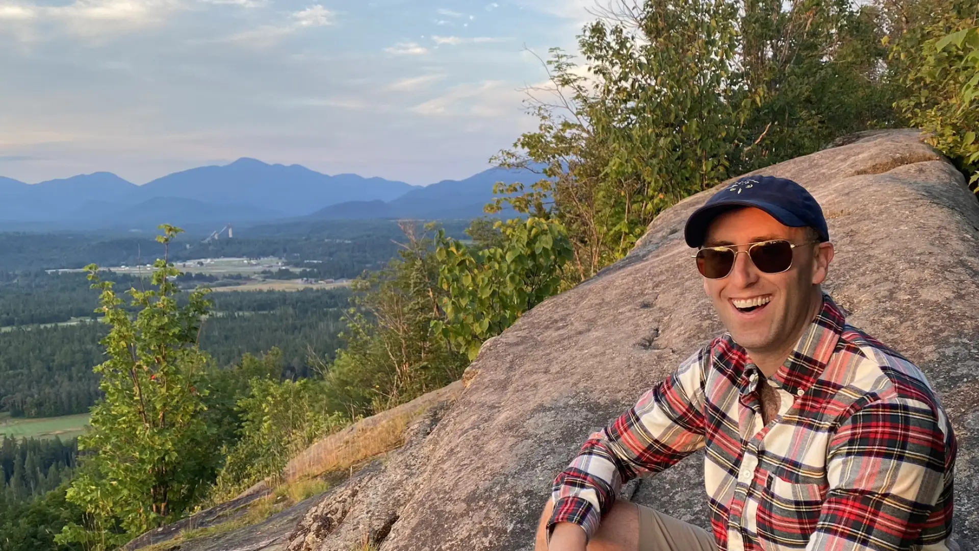 A bare foot man in sunglasses and a flannel sits atop a rocky mountain top.