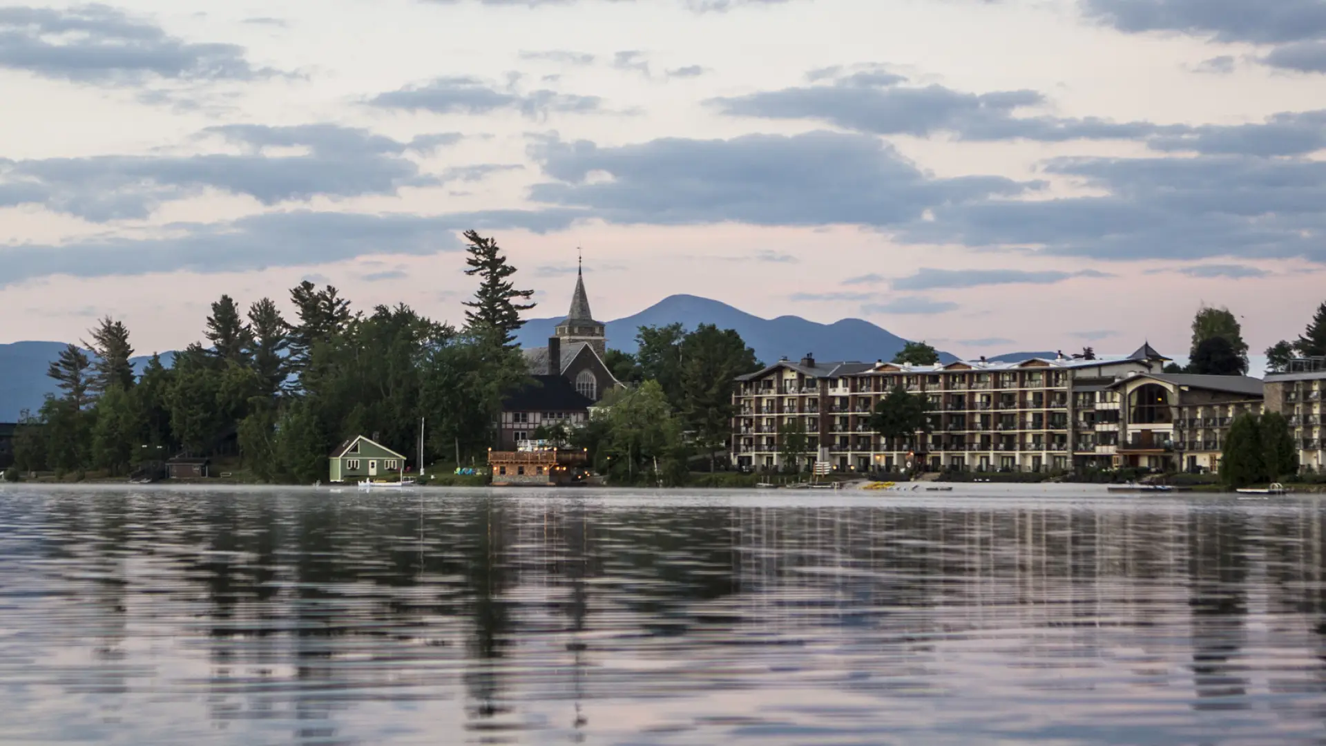 A wide hotel, church, and other buildings amid trees and a backdrop of mountains with a lake in the foreground.